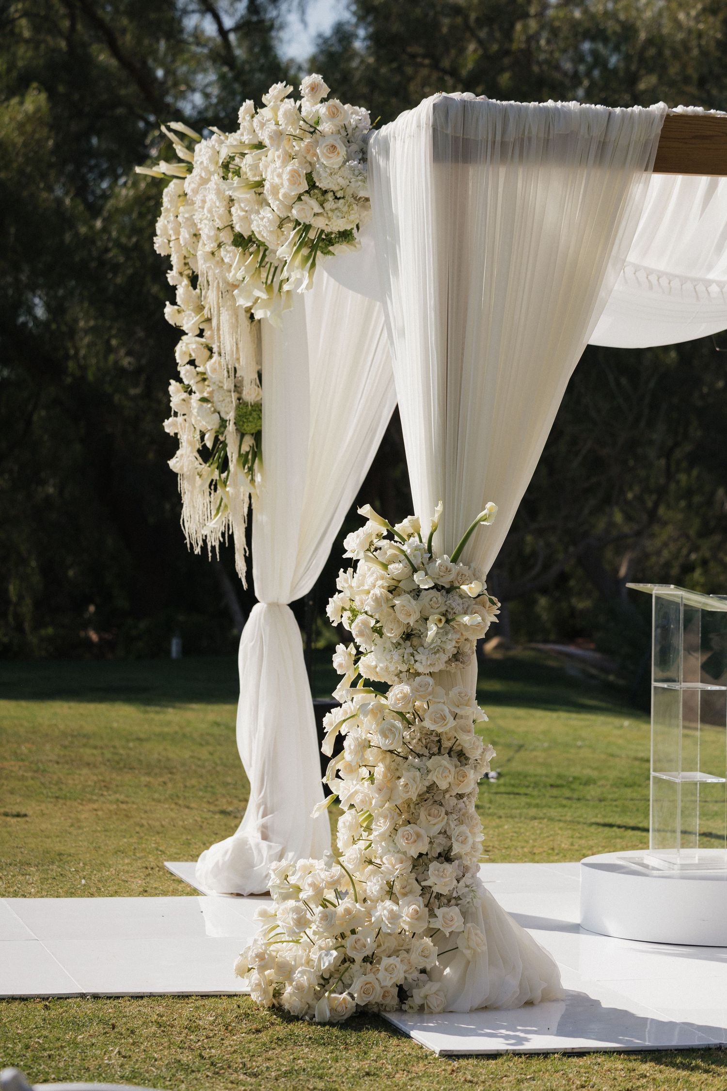 Elegant wedding arch adorned with cascading white roses and floral arrangements, draped with flowing white fabric outdoors.