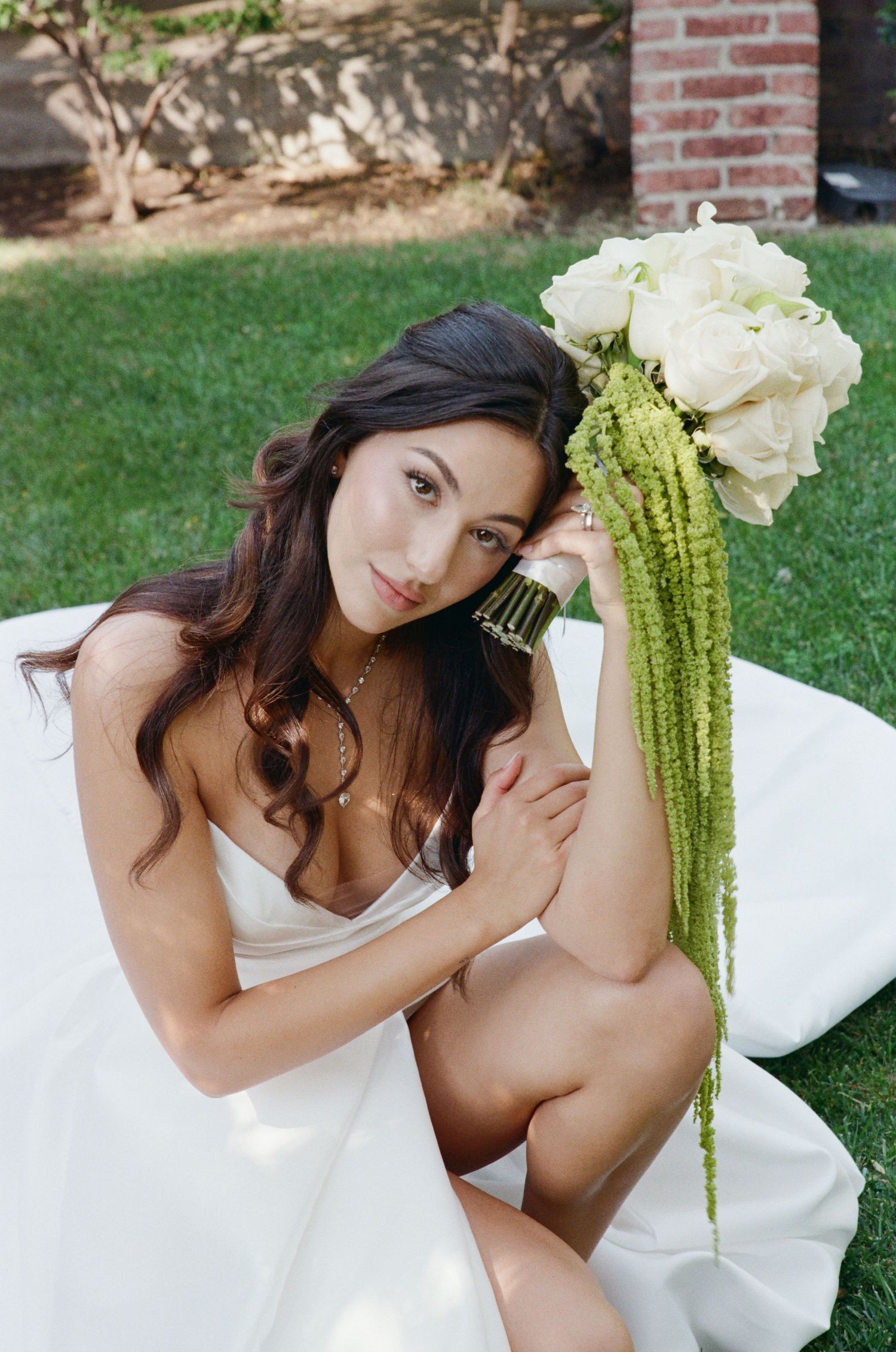 Bride in white gown sitting on grass holding elegant bouquet of white roses with green amaranthus.