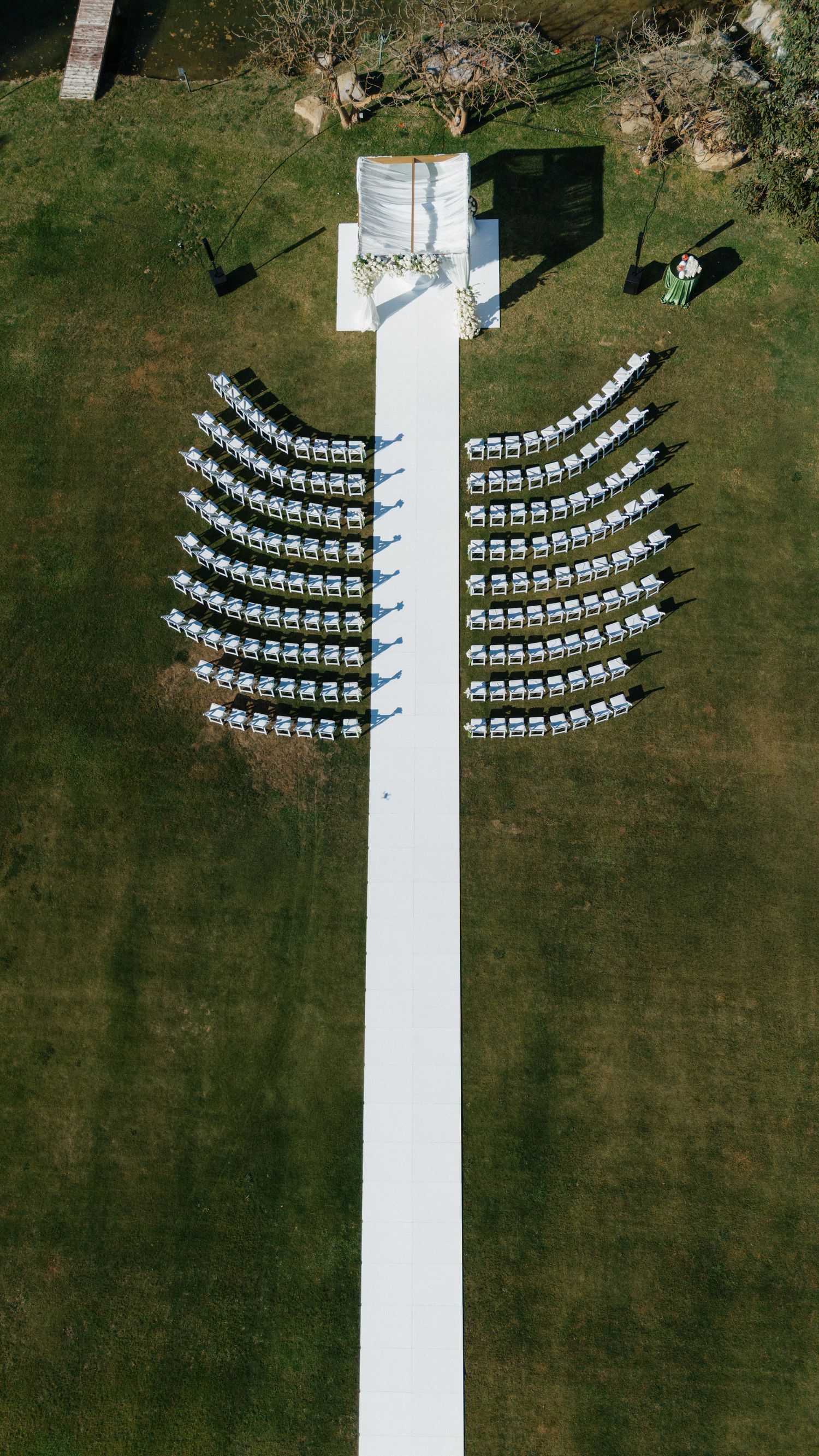 Aerial view of outdoor wedding ceremony setup with curved rows of white chairs and long white aisle on green lawn.