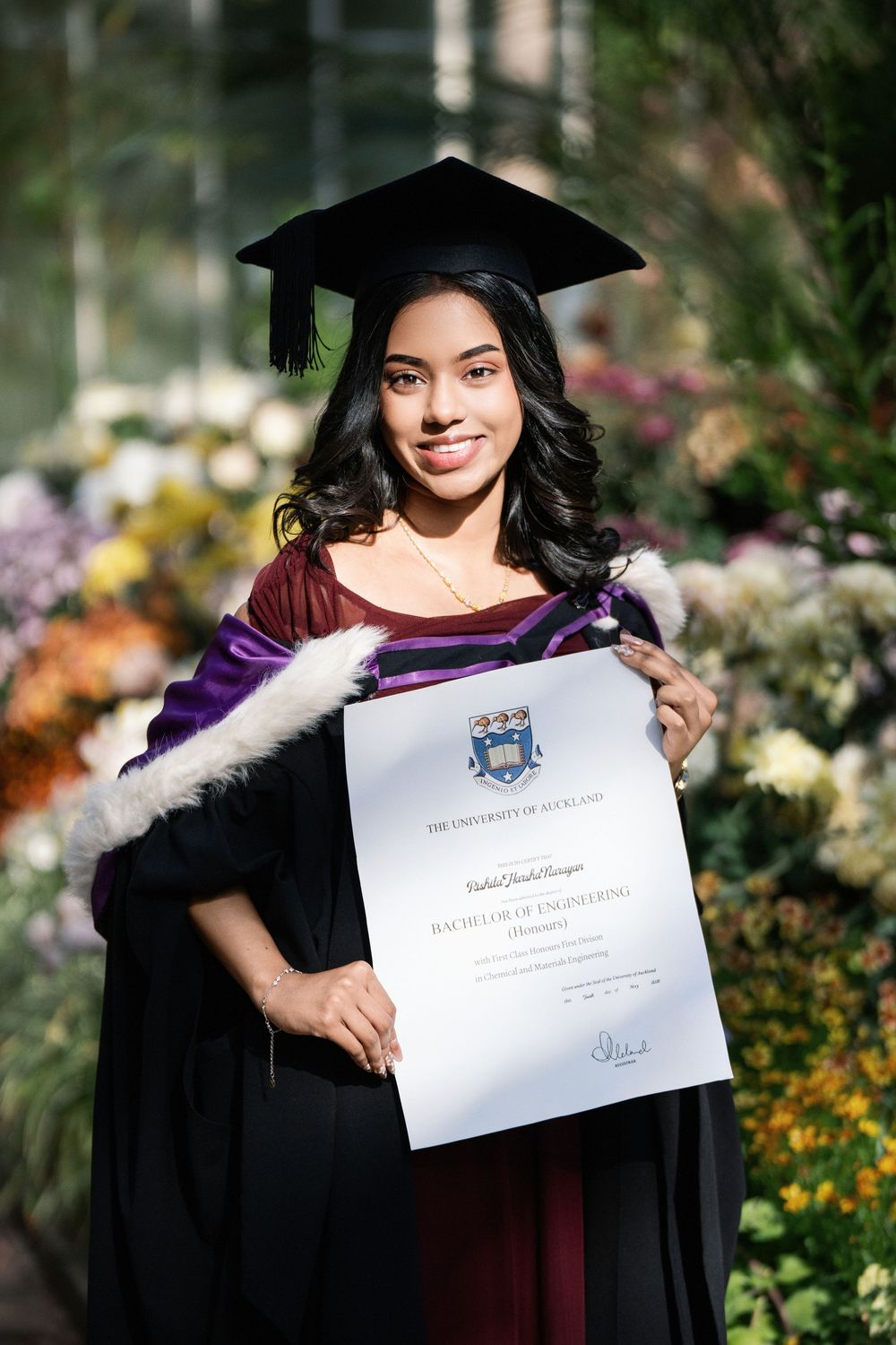 Elegant indoor graduation photo of a student holding her diploma amidst the lush floral displays of the Winter Garden, Auckland Domain.