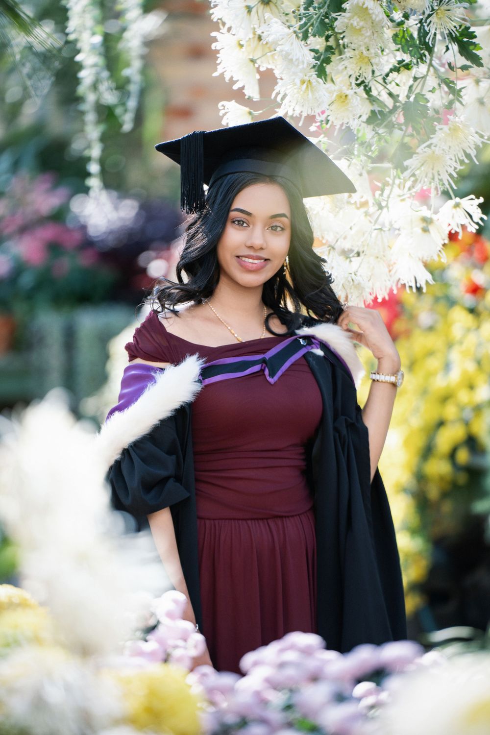 Beautiful graduation photos of a student in a maroon gown surrounded by blooming flowers inside the Winter Garden at Auckland Domain.