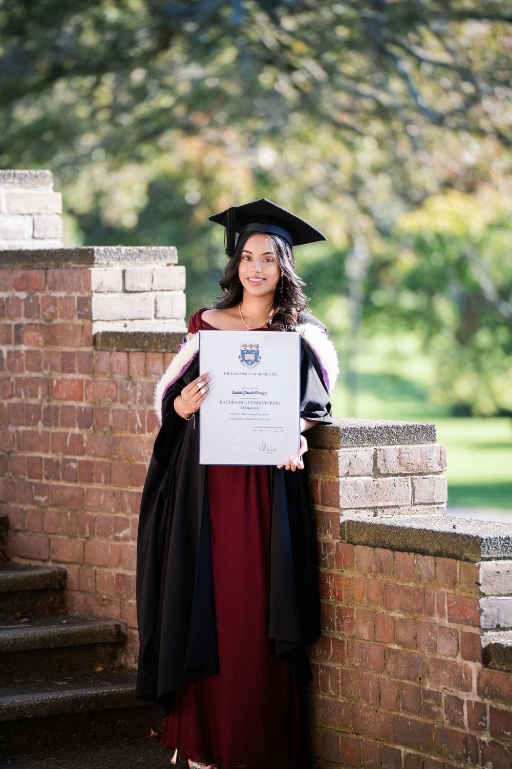 A graduate standing on brick stairs holding her degree in Auckland Domain for outdoor graduation photos.