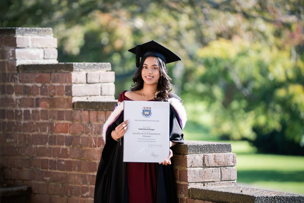 A graduate in a maroon gown holding her diploma proudly near historic brick architecture in Auckland Domain.
