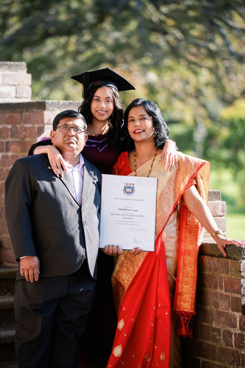Family graduation photo featuring a graduate and her parents standing by a brick wall in Auckland Domain.