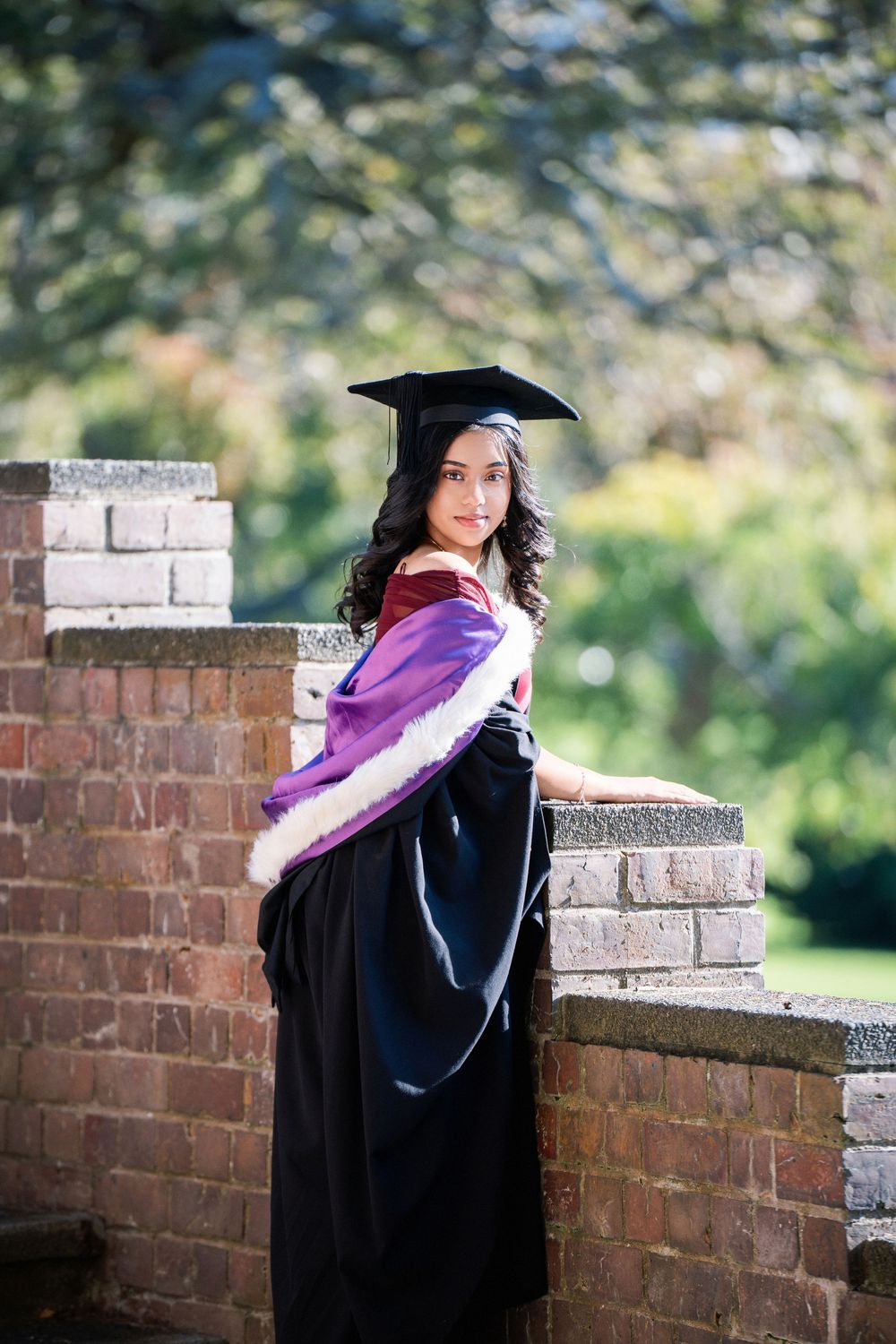 A smiling graduate standing on a brick wall with a scenic background in Auckland Domain for her graduation photos.