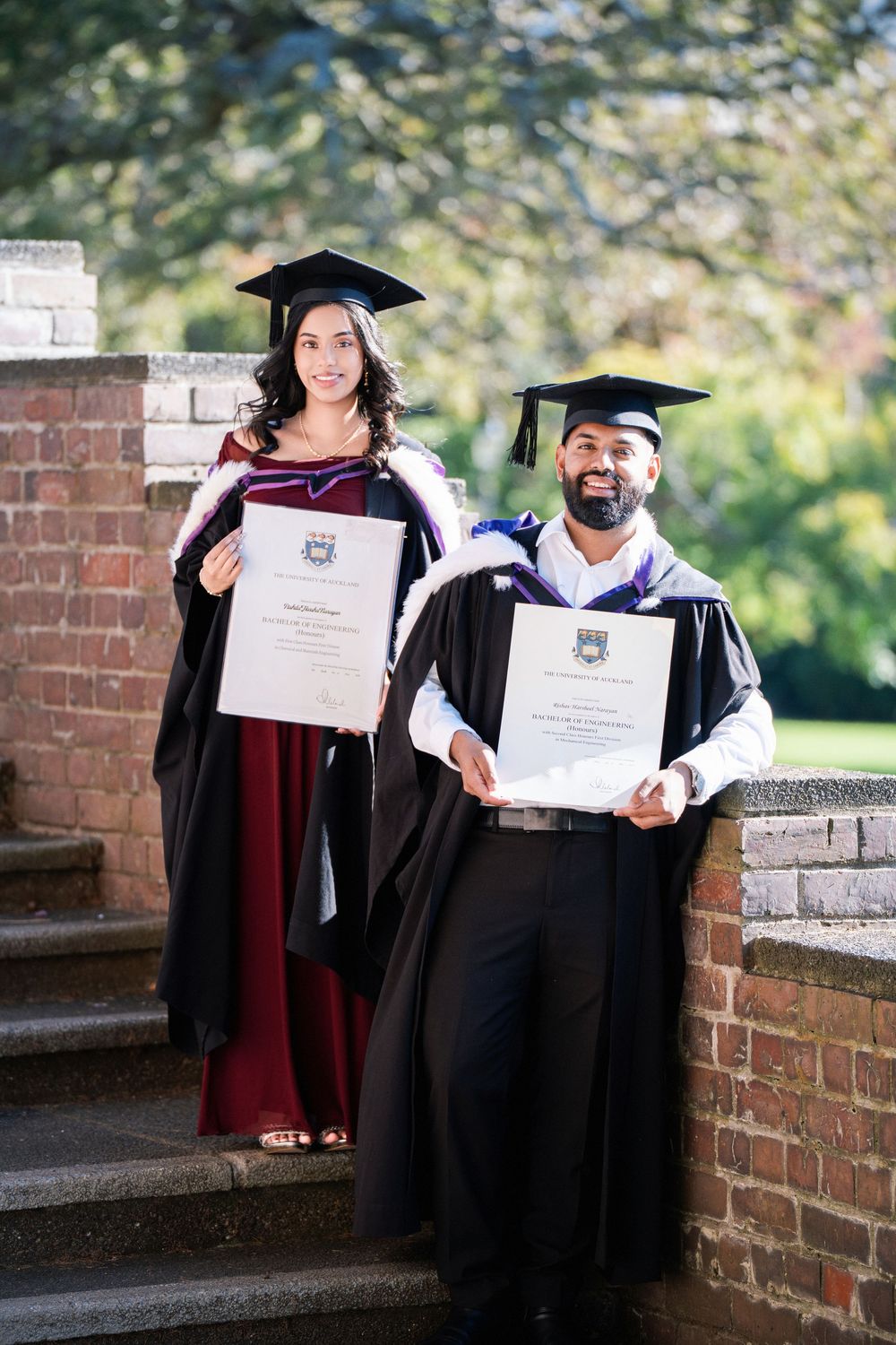 A graduate and her partner holding degrees on historic brick steps near the Winter Garden, Auckland Domain for graduation photos.