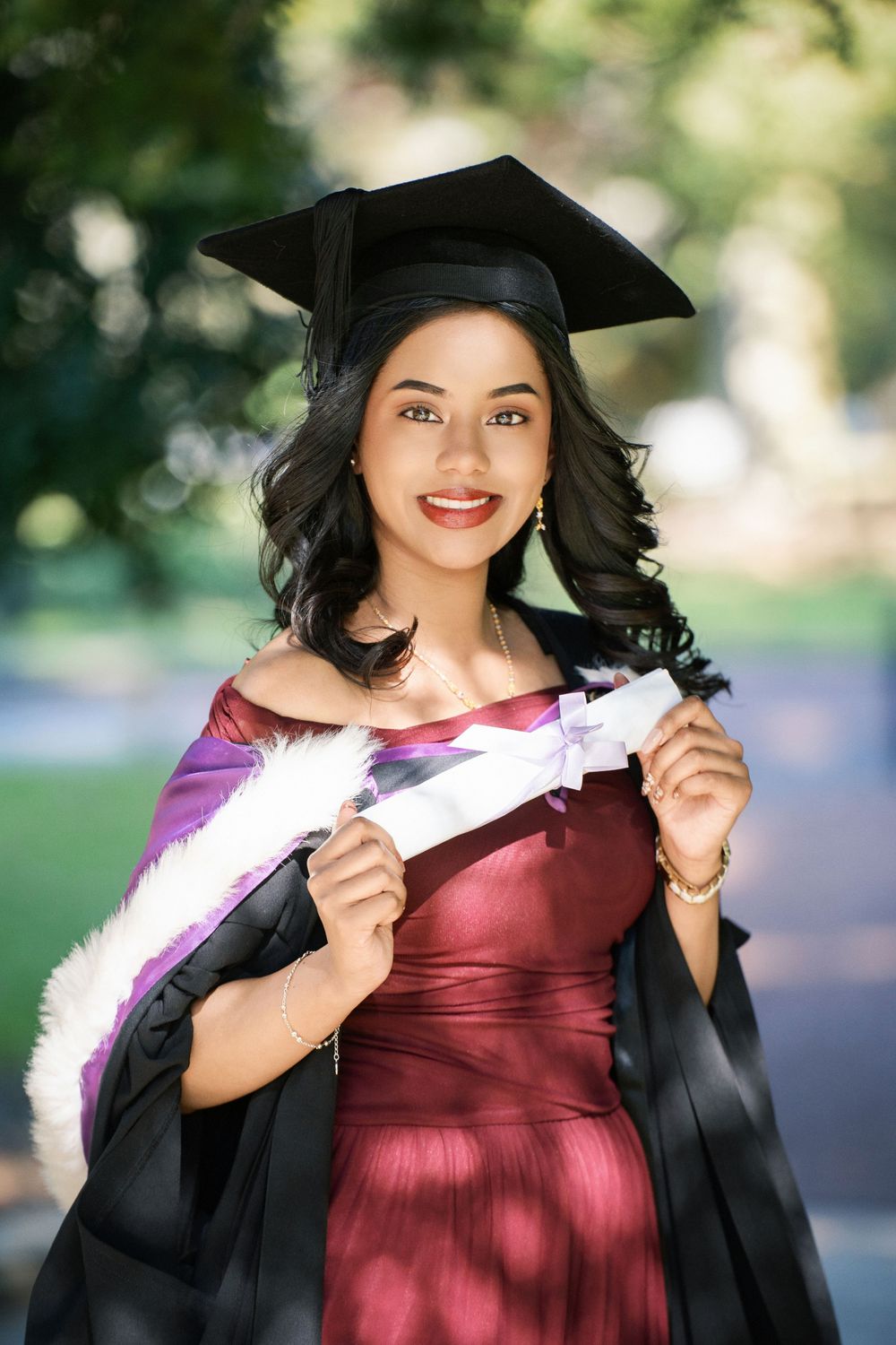 Close-up portrait of a proud graduate in a maroon gown holding her degree in Auckland Domain.