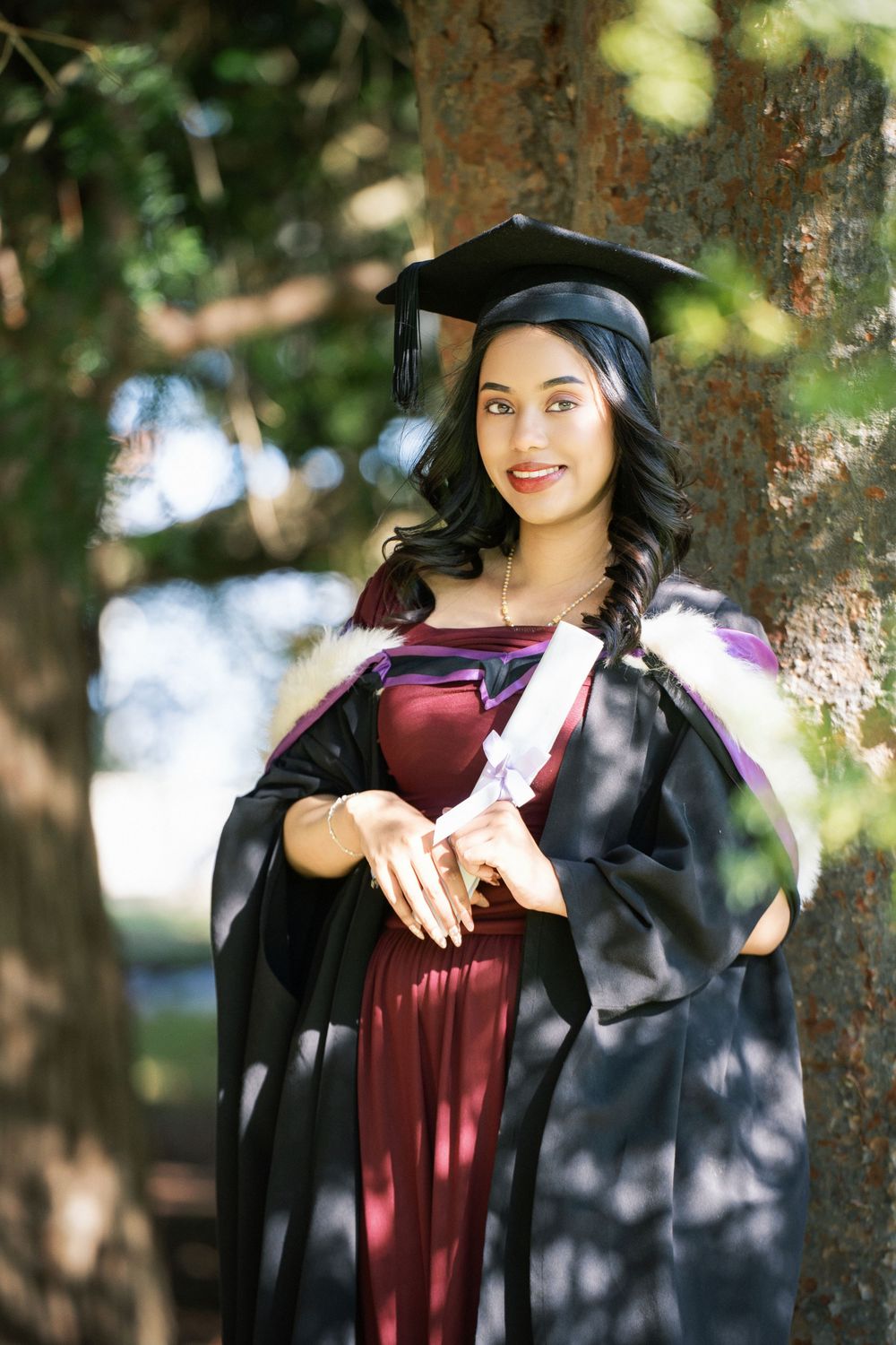 A graduate in a maroon gown leaning against a large tree in Auckland Domain for her graduation photos.