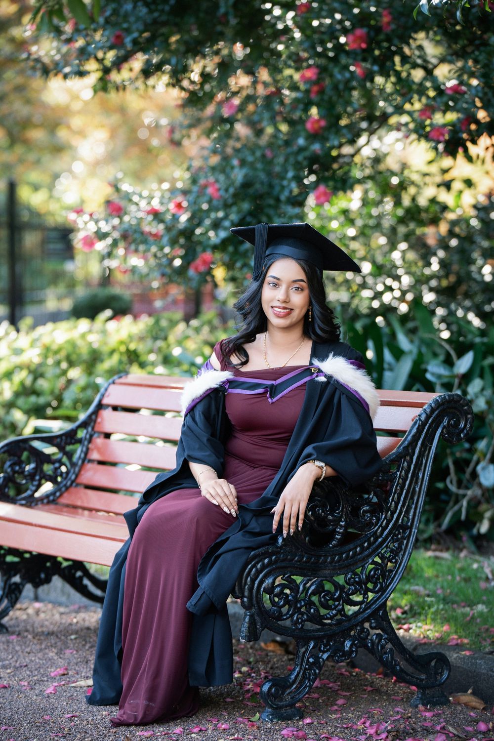 A beautiful graduate in a maroon gown sitting on a park bench surrounded by greenery in Auckland Domain.