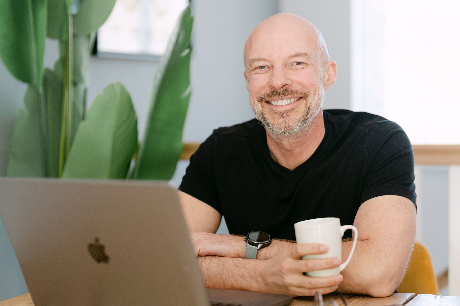 Smiling bald man with beard holding coffee mug working on Apple MacBook laptop in bright modern office.