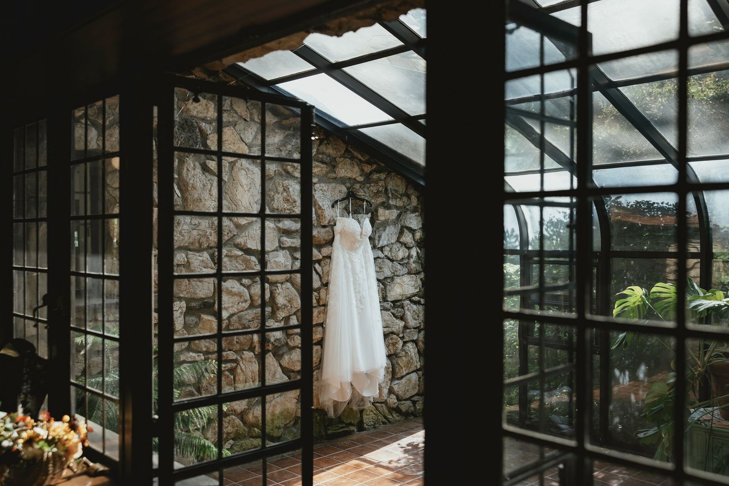 A white wedding dress hanging in a rustic glass greenhouse with stone walls and lush greenery visible outside.