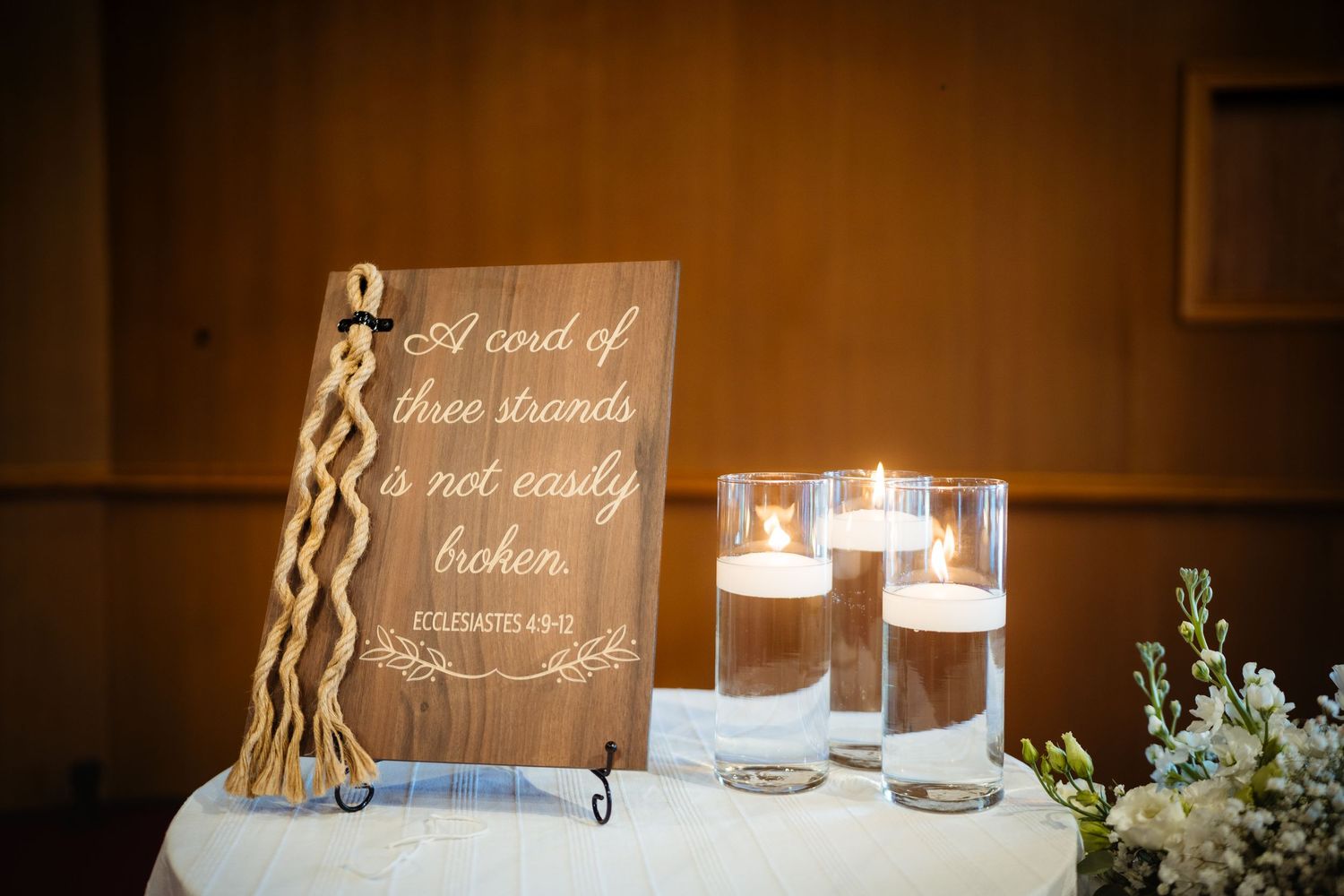 Wooden wedding sign reading 'A cord of three strands is not easily broken' with braided rope and floating candles.