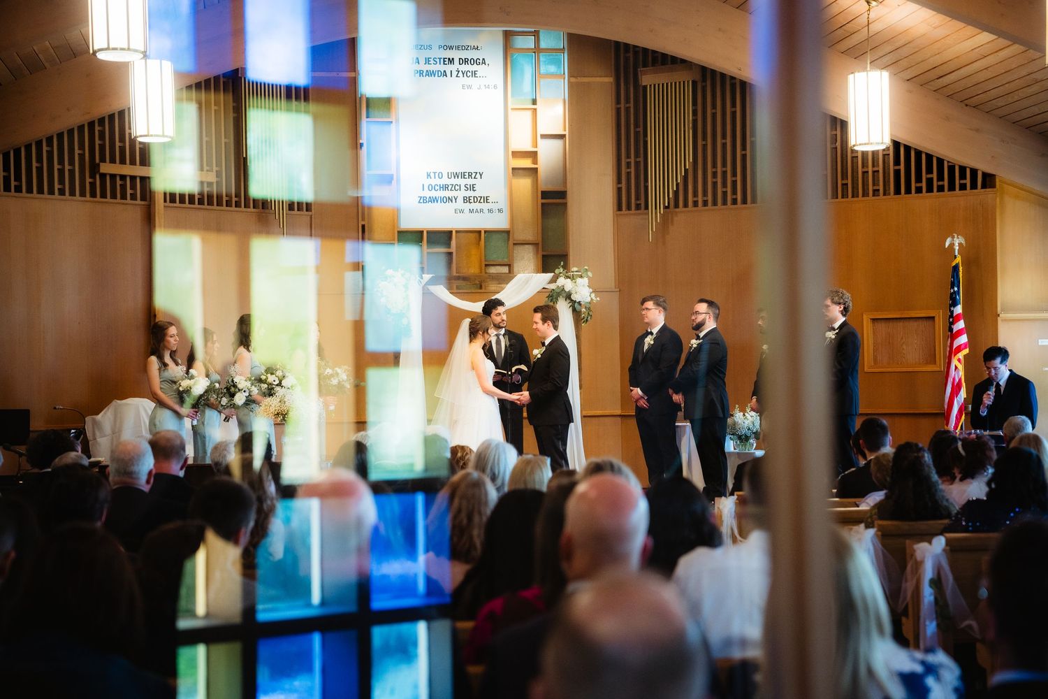 Wedding ceremony in a modern church with colorful stained glass windows, bride and groom at the altar with guests seated.
