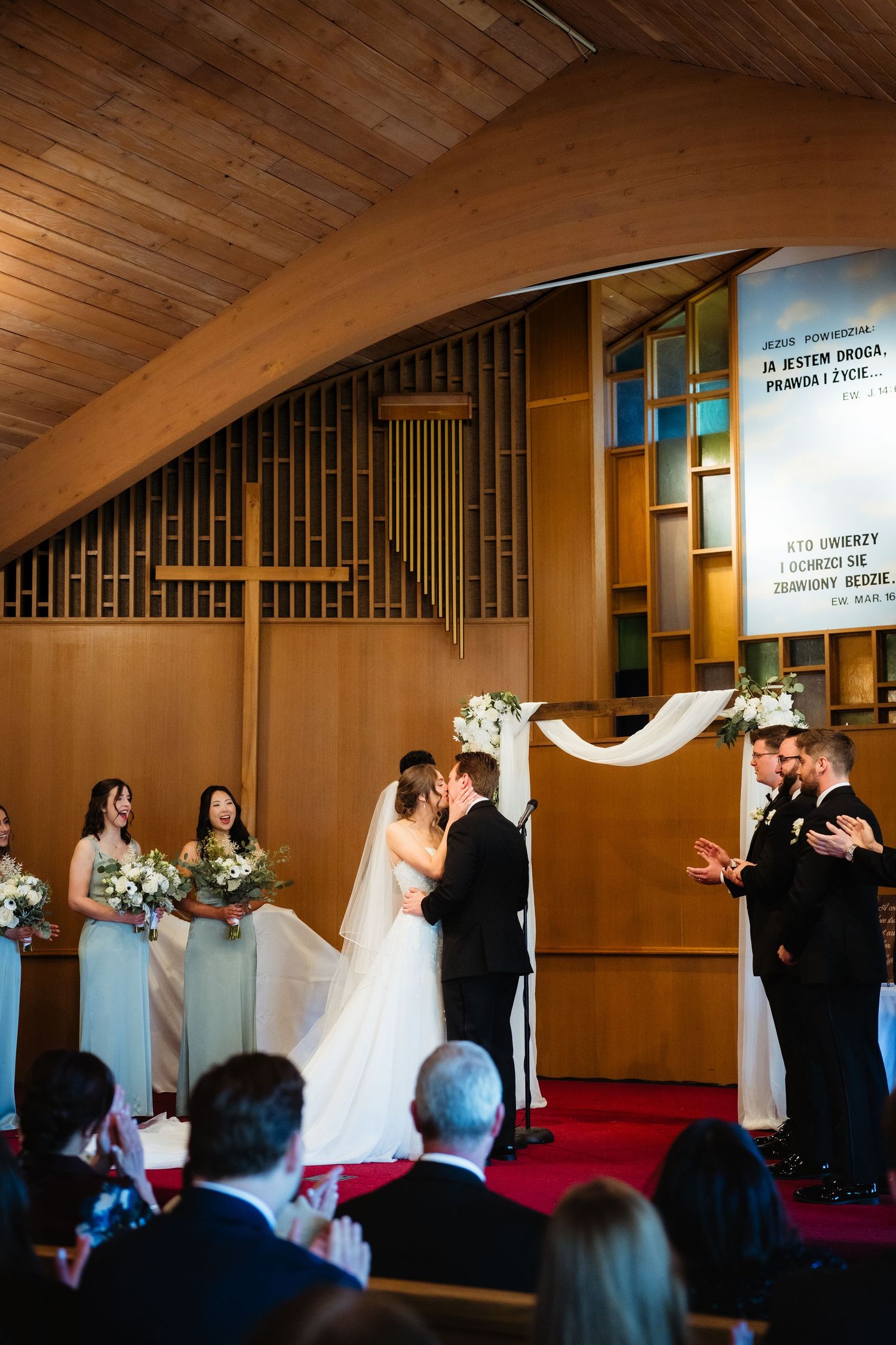 Bride and groom share first kiss at church altar surrounded by bridesmaids and groomsmen during wedding ceremony.