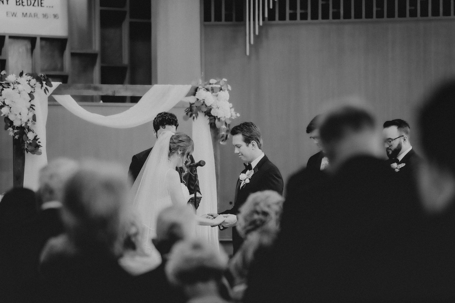 Black and white wedding ceremony photo of bride and groom at altar with officiant in elegant church setting.
