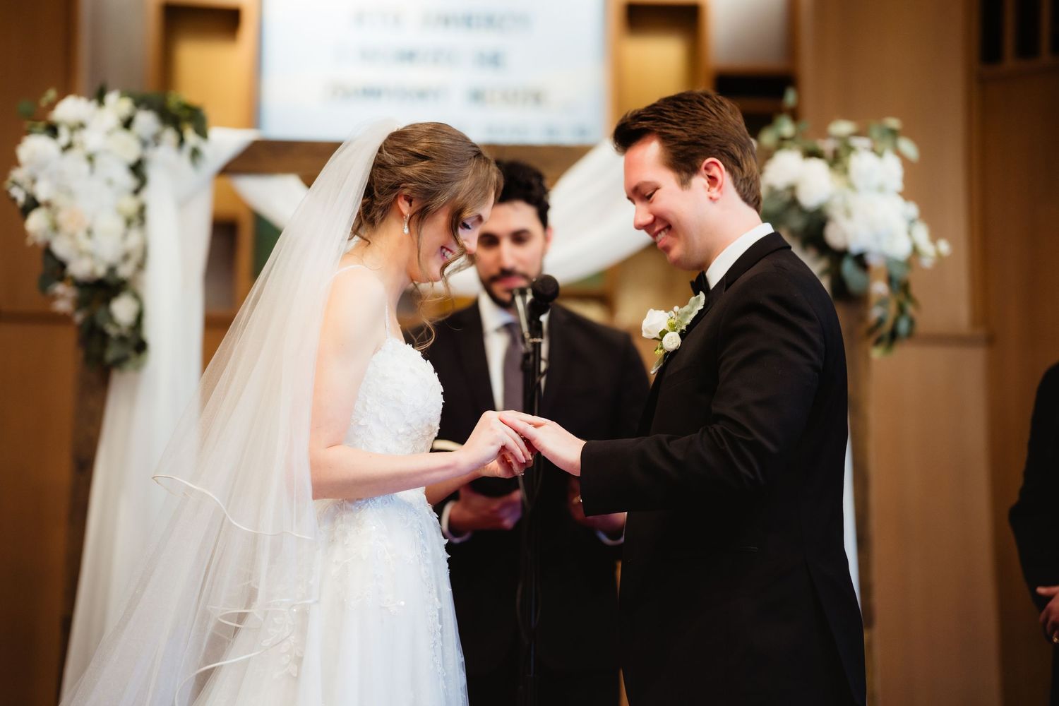 Bride placing wedding ring on groom's finger during ceremony, officiant standing behind them, white floral decorations.