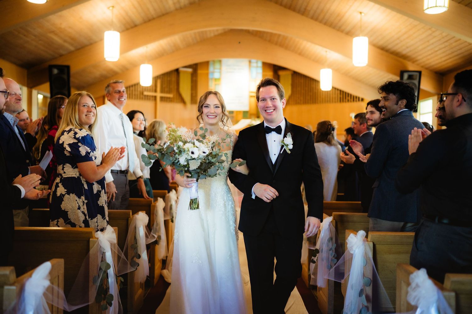 Bride and groom walk down church aisle after wedding ceremony, smiling guests applauding in wooden pew chapel.