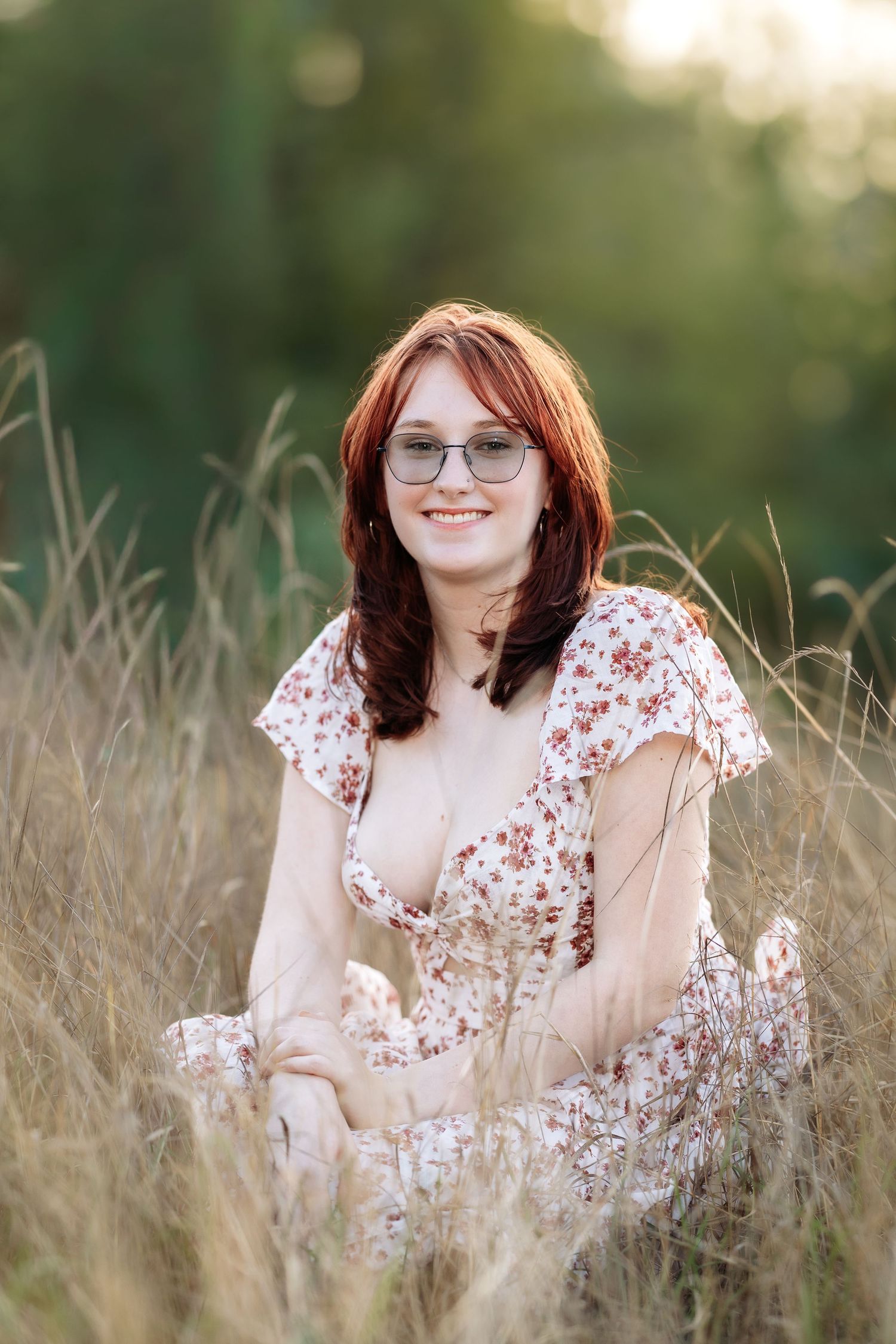 Young redhead woman with glasses sitting in tall golden grass, wearing a floral dress, smiling outdoors at sunset.