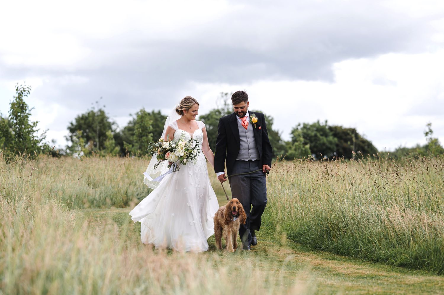 Bride and groom walking with their dog through a lush green meadow path on their wedding day at Oxleaze Barn.