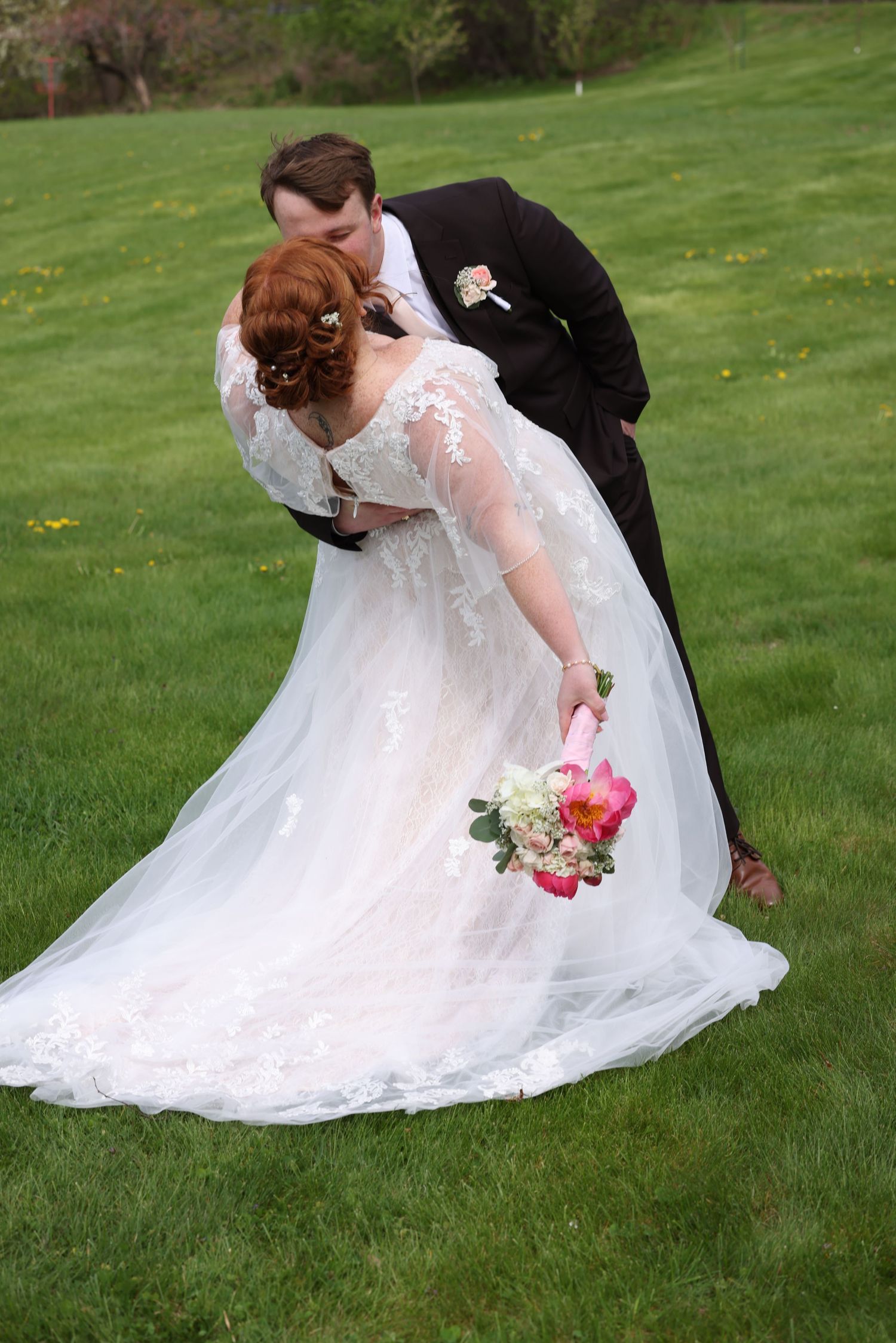 Bride and groom sharing a romantic dip kiss on green lawn, bride holding pink and white bouquet.