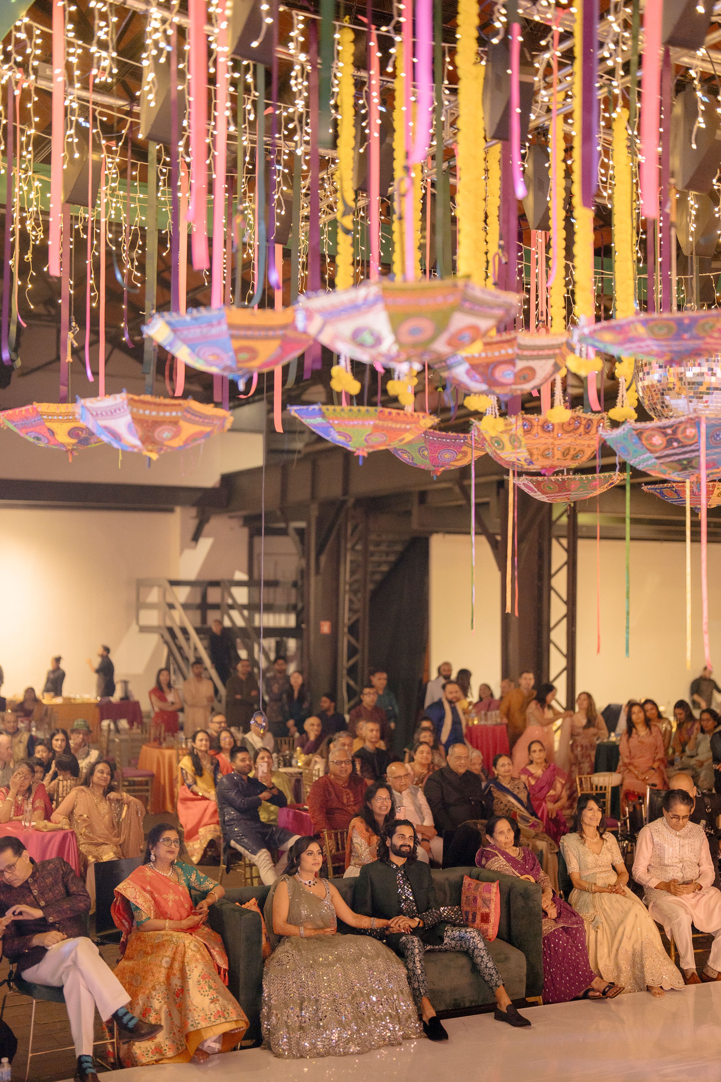 Colorful Indian wedding celebration with guests in traditional attire under vibrant hanging umbrellas and ribbon decorations.