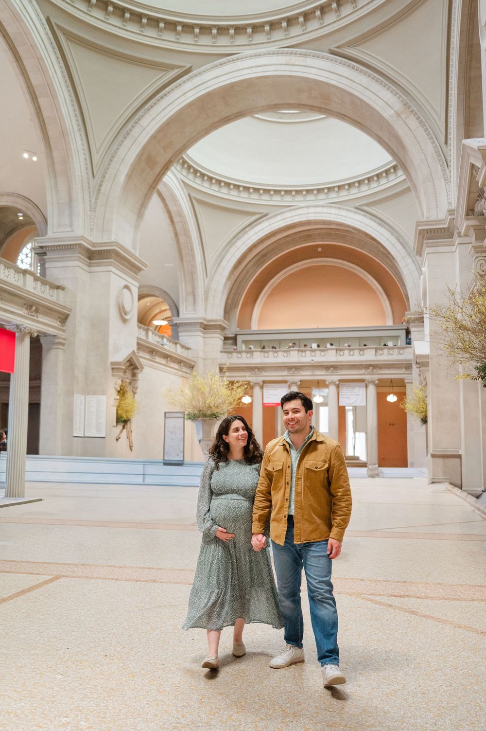 Pregnant couple holding hands in grand museum hall with arched ceilings, woman in sage dress, man in tan jacket.
