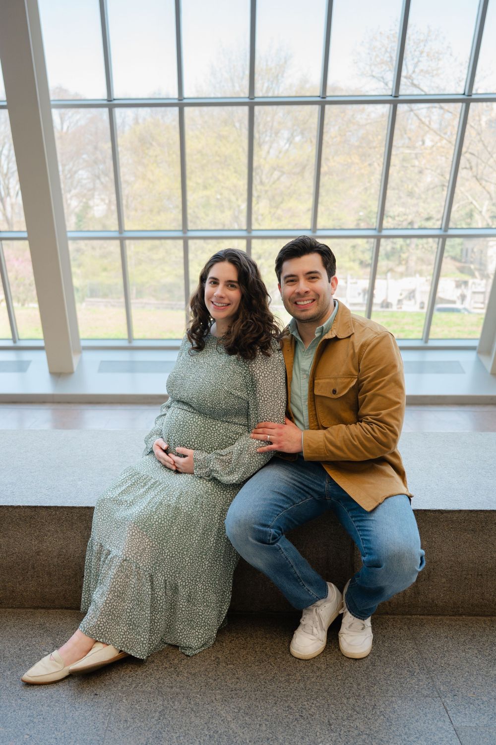 Expecting couple posing by large window, woman in floral sage dress cradling baby bump, man in tan jacket smiling.