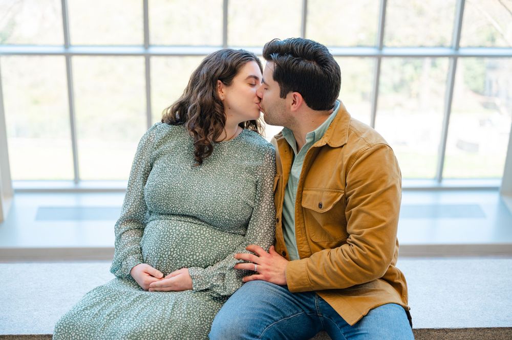 Pregnant couple sharing a tender kiss while sitting together near large windows in a maternity photo session.