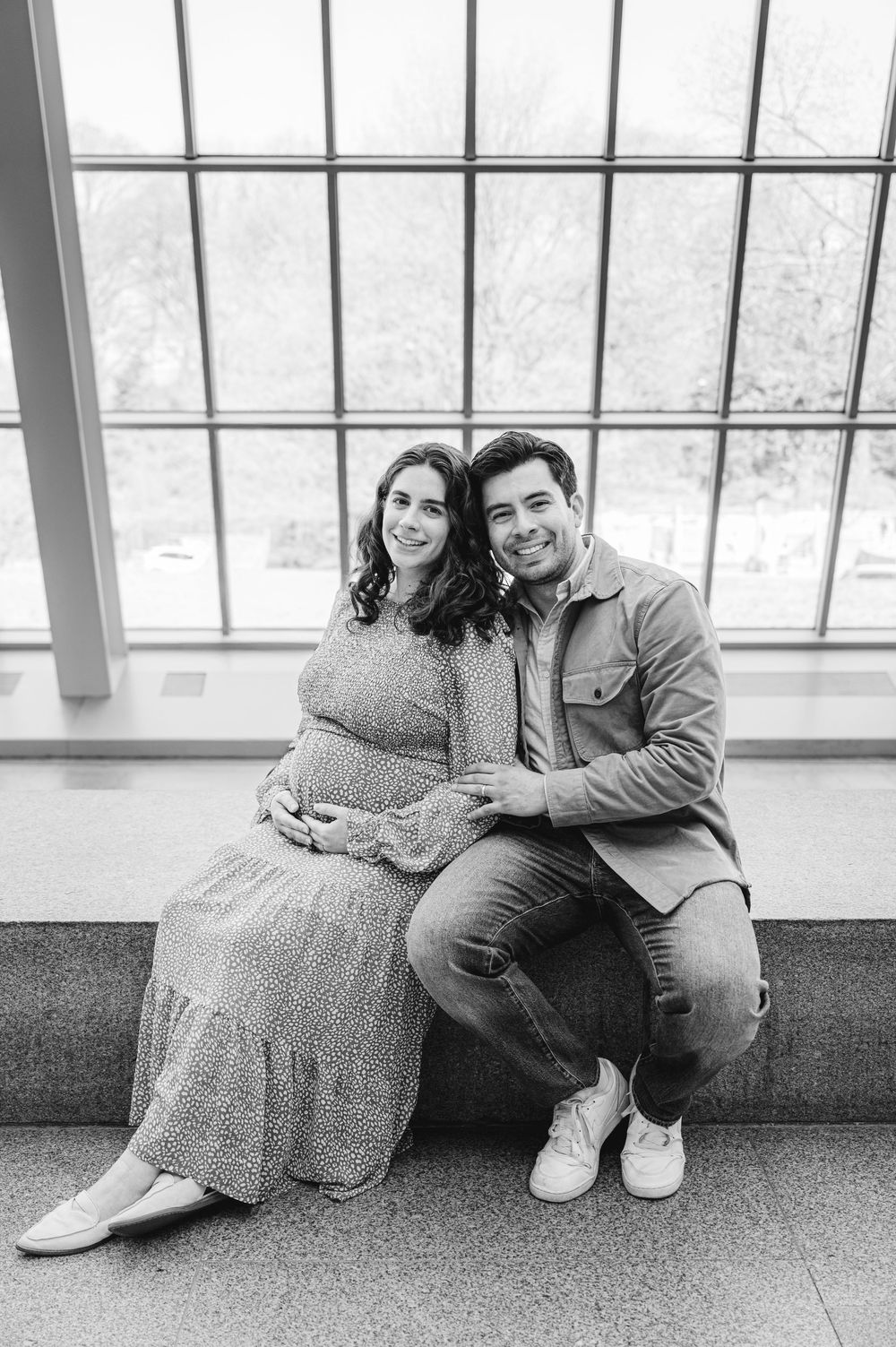 Black and white maternity photo of a smiling couple sitting together, the woman in a floral dress cradling her baby bump.