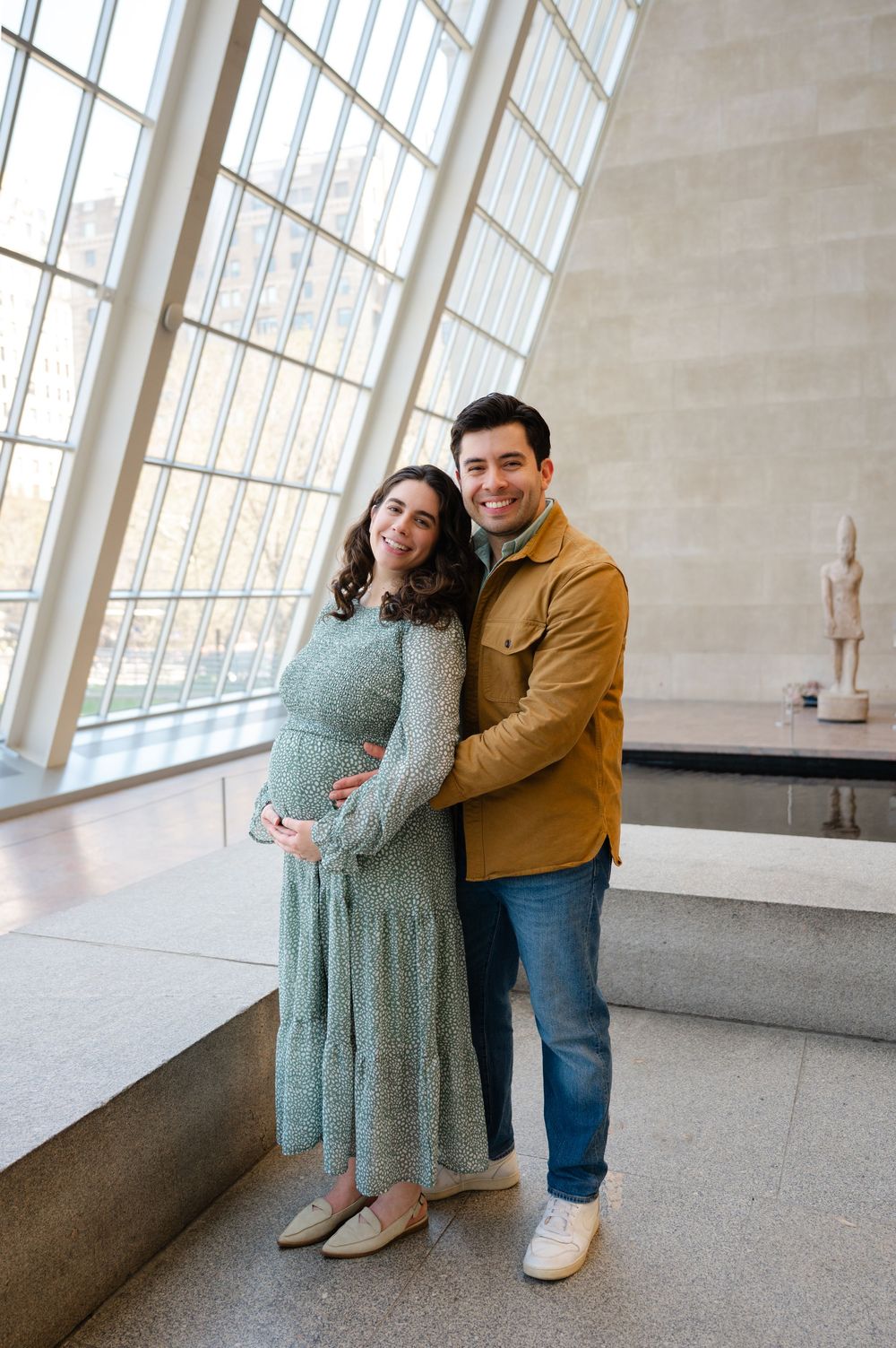 Smiling pregnant couple embraces near tall windows inside a bright museum gallery.