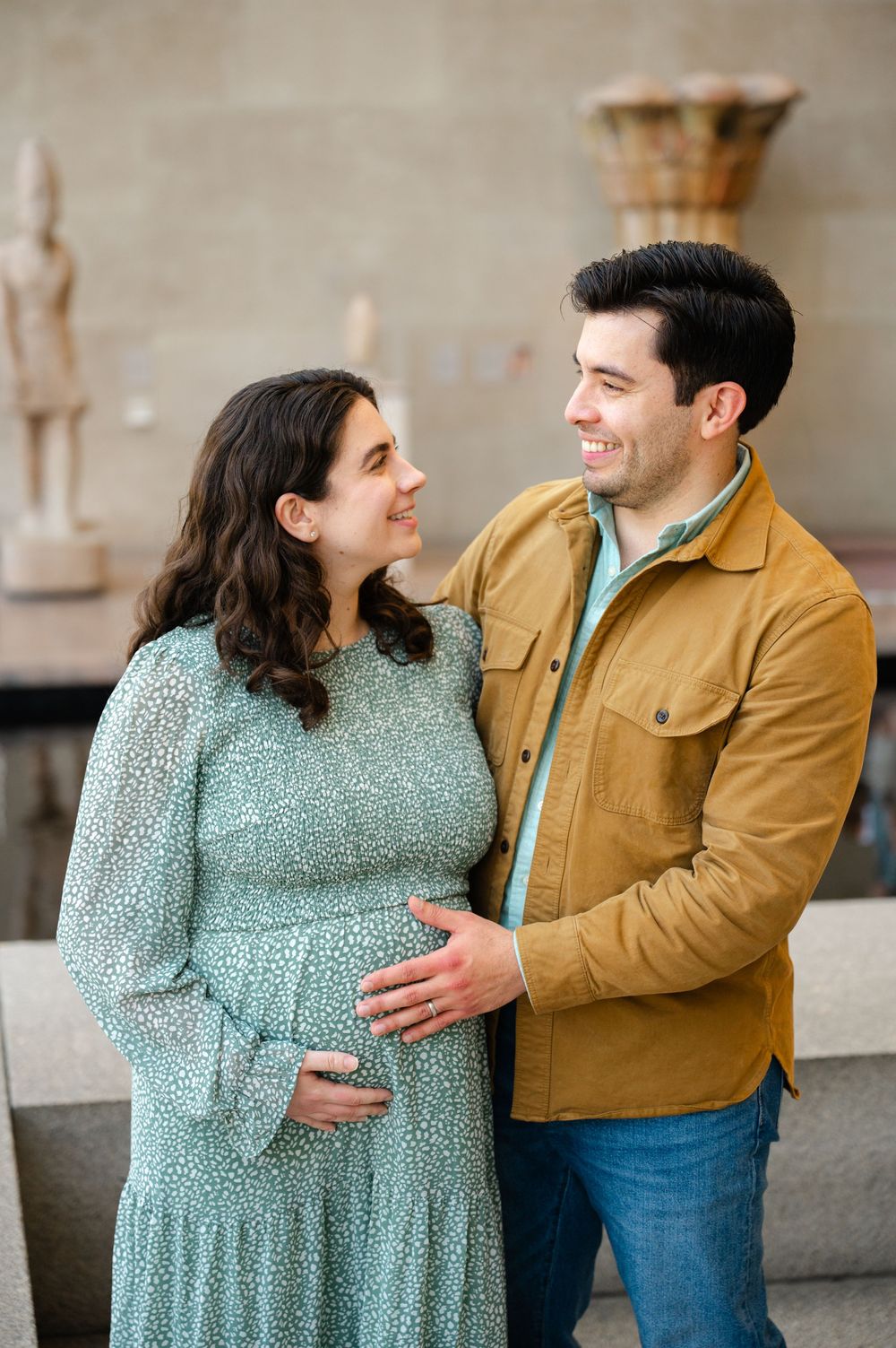 Smiling expectant couple gazing at each other lovingly, man's hand on pregnant belly, surrounded by museum artifacts.