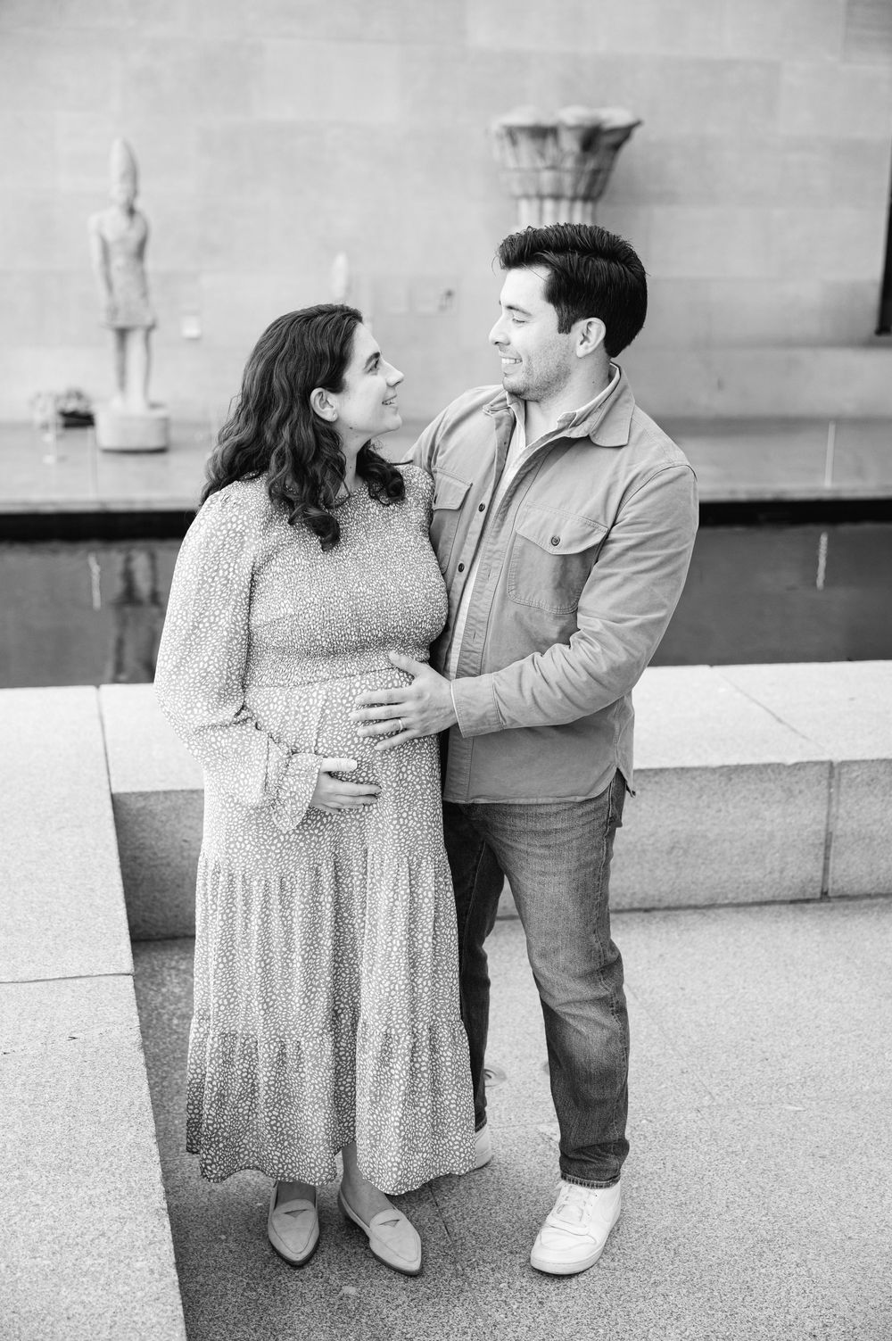 Black and white maternity photo of a couple smiling at each other, man gently holding pregnant woman's belly outdoors.