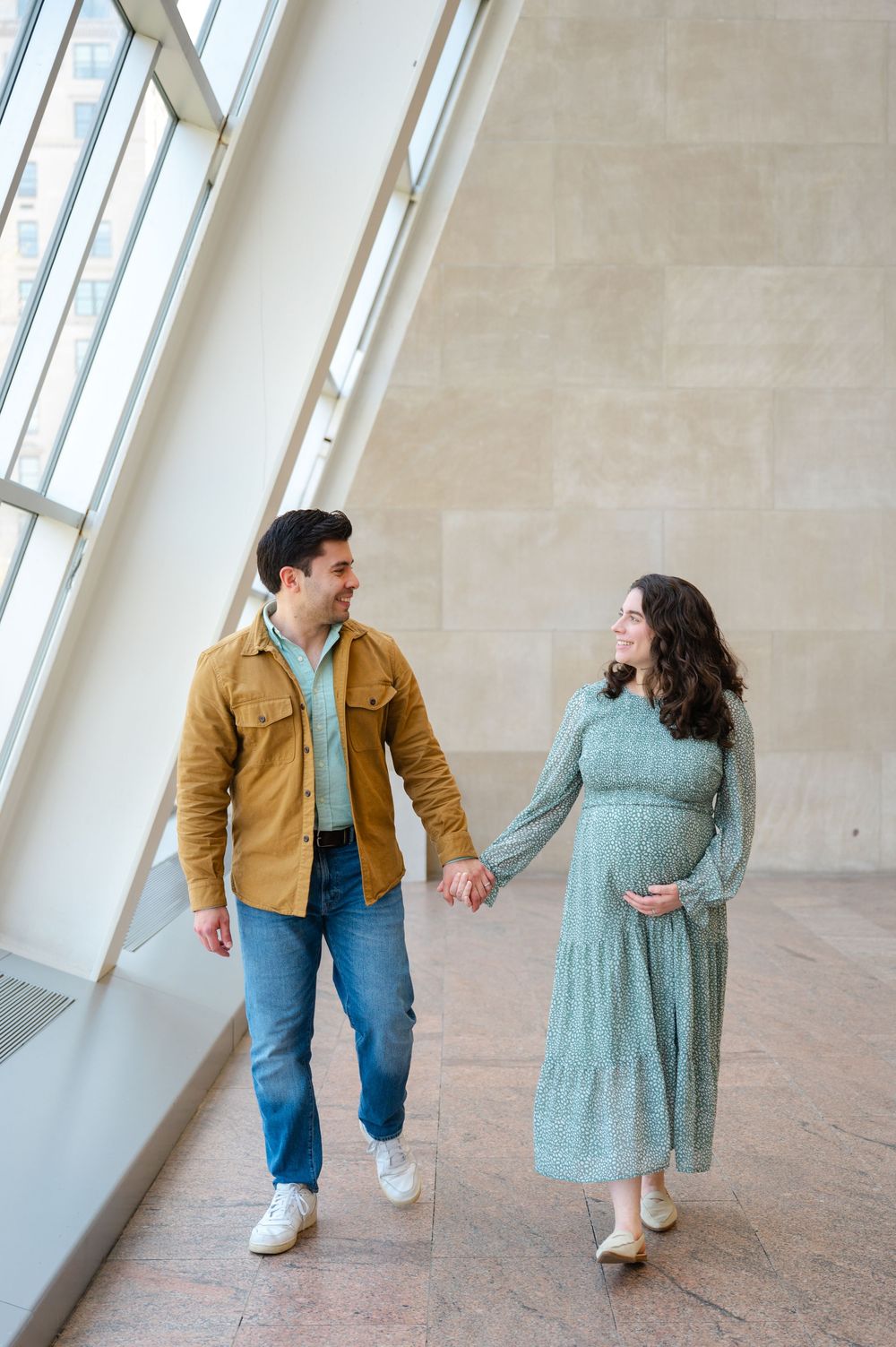Expecting couple holds hands and smiles at each other while walking through a sunlit museum hallway.