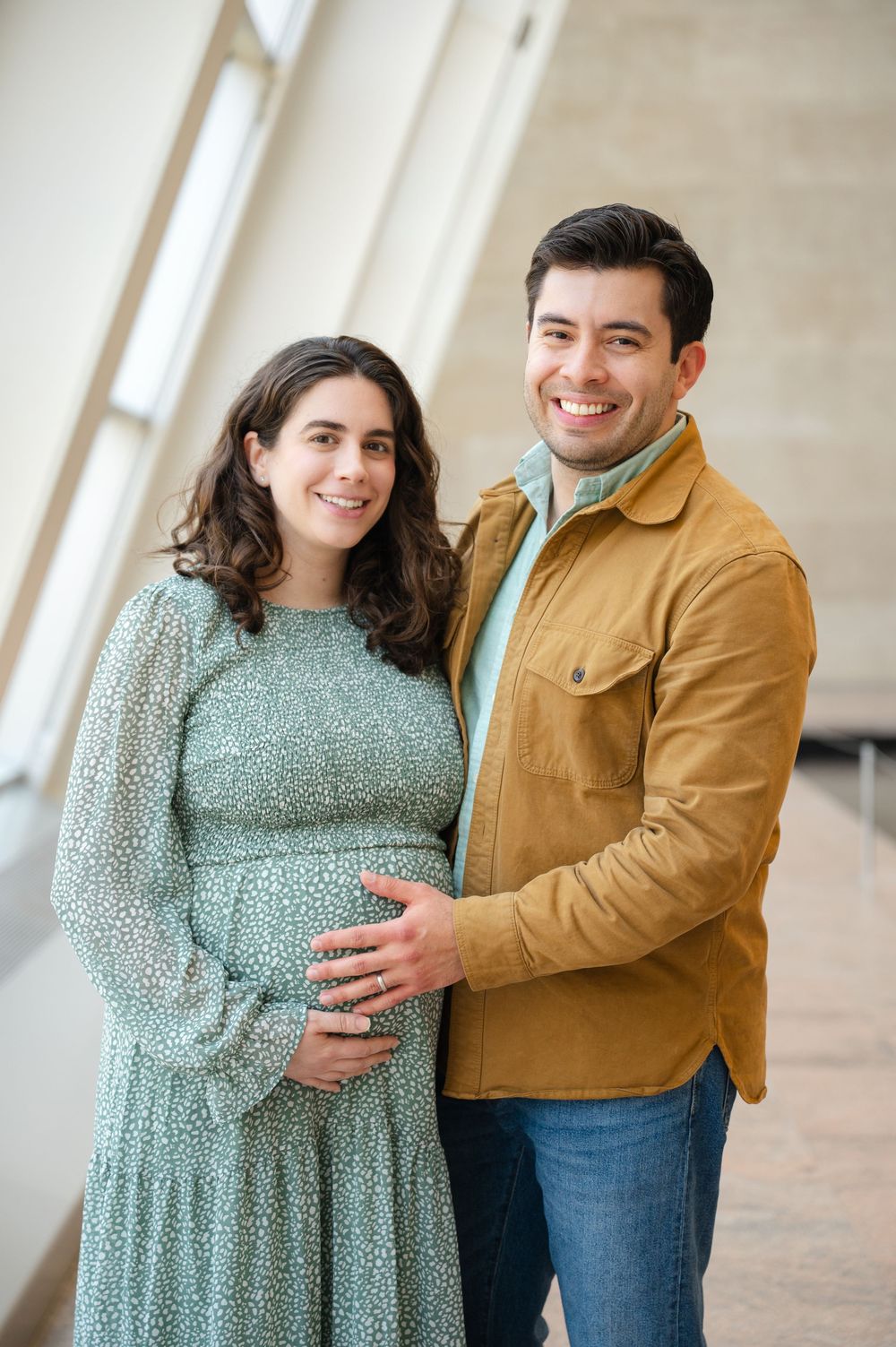 Happy couple posing for maternity photo, woman in floral dress cradling baby bump, man in tan jacket smiling.