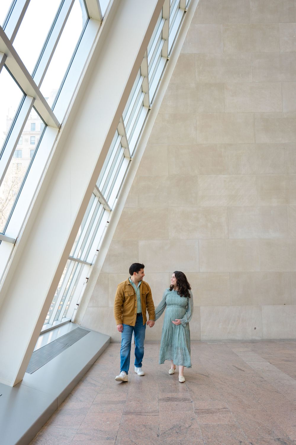 Expecting couple holds hands walking through sunlit museum atrium with large windows.