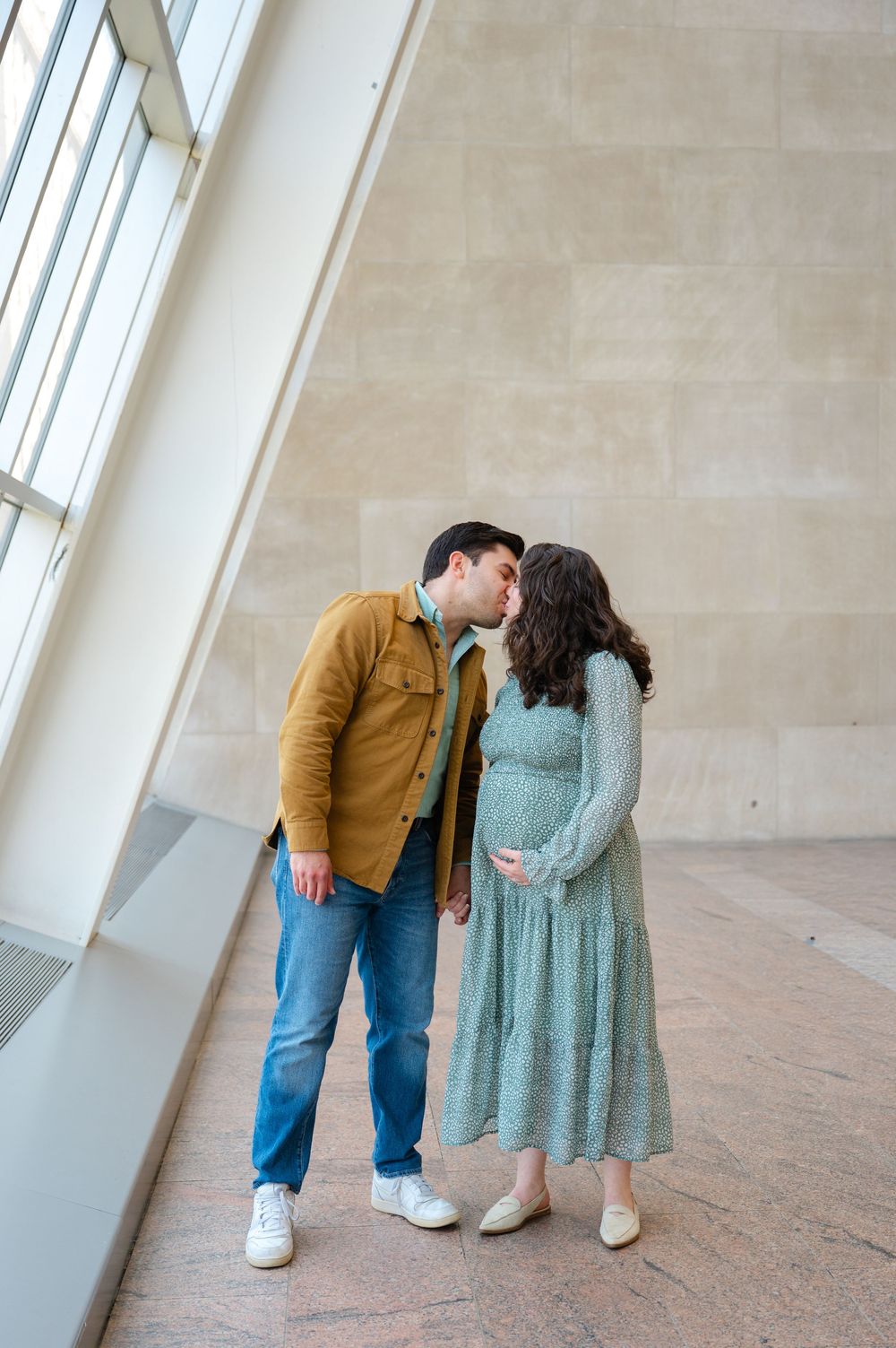 Pregnant couple sharing a tender kiss in a sunlit museum atrium, woman in sage floral dress cradling her baby bump.