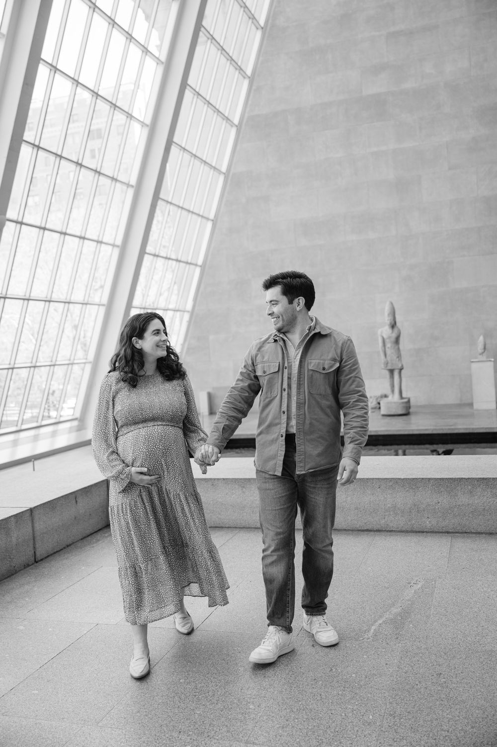 Black and white photo of a pregnant couple holding hands and smiling in a museum gallery.