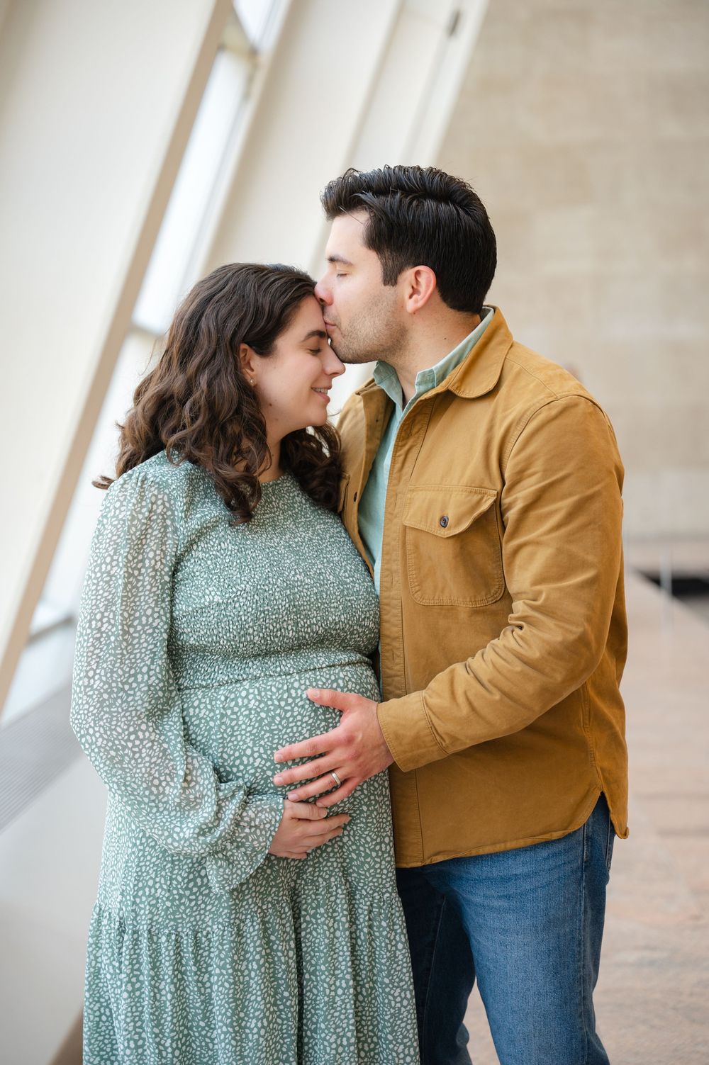 Expectant couple kissing intimately as man gently holds pregnant woman's baby bump in a bright museum setting.