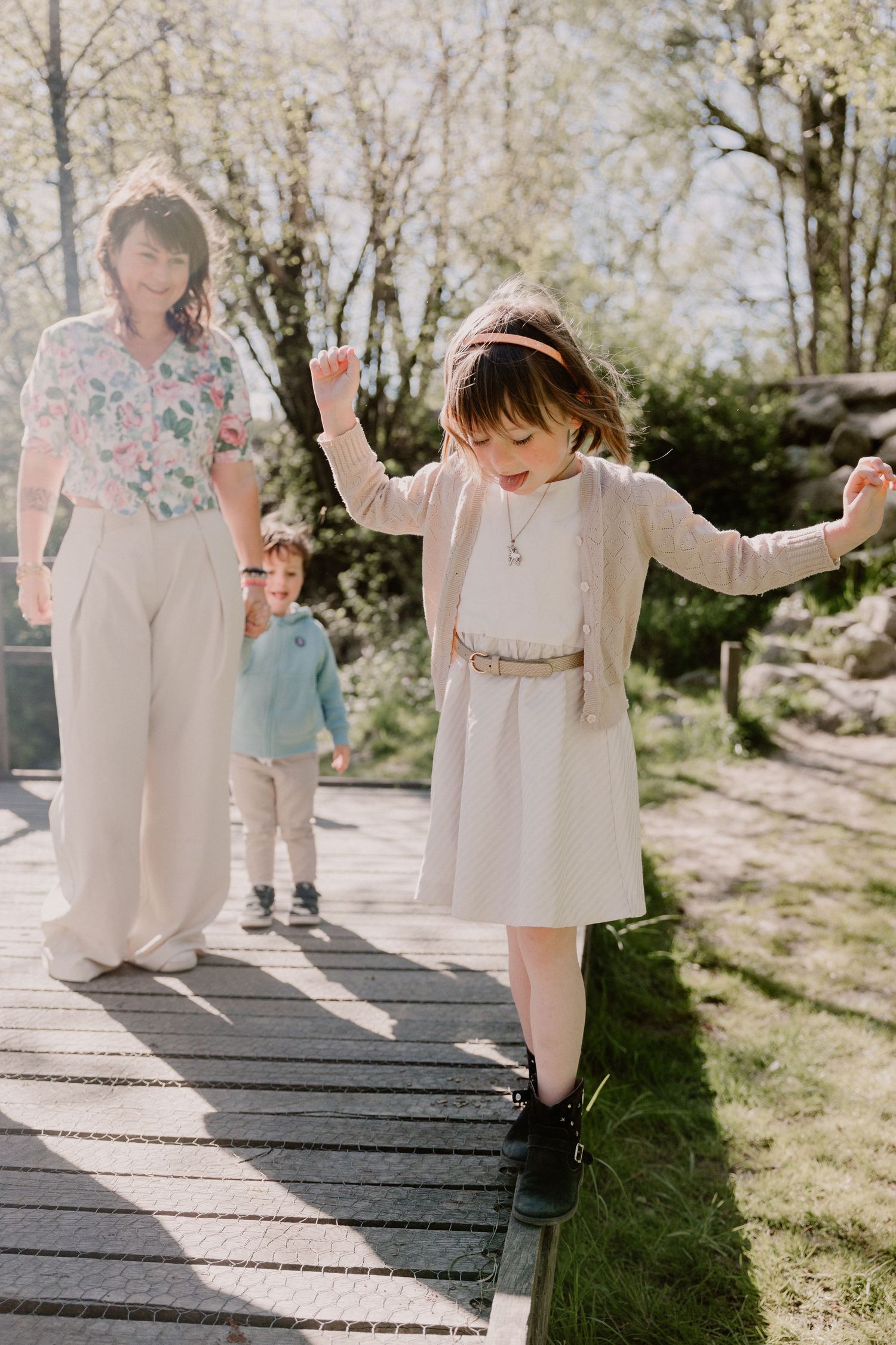 Young girl in beige dress balancing on wooden boardwalk while mother and brother walk behind.