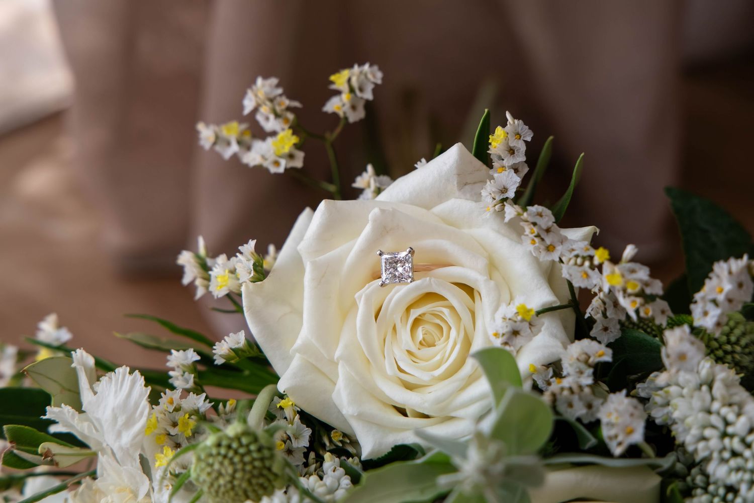 Close-up of a white rose bouquet with small wildflowers and a diamond engagement ring nestled among the blooms.