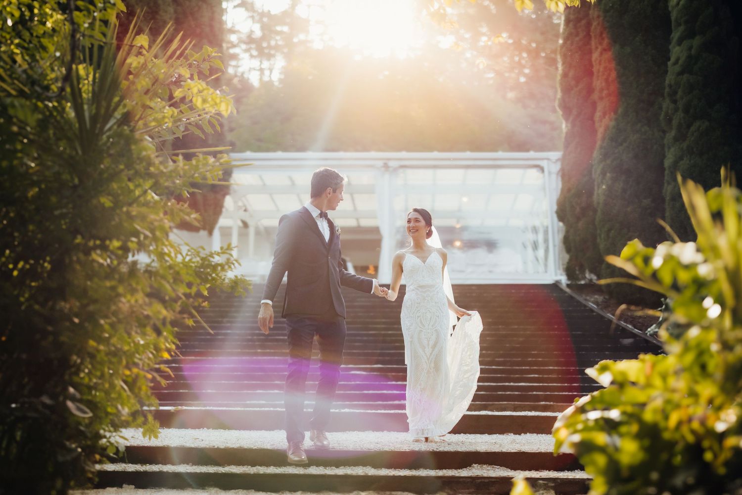 Wedding couple ascending sunlit garden stairs surrounded by lush greenery.