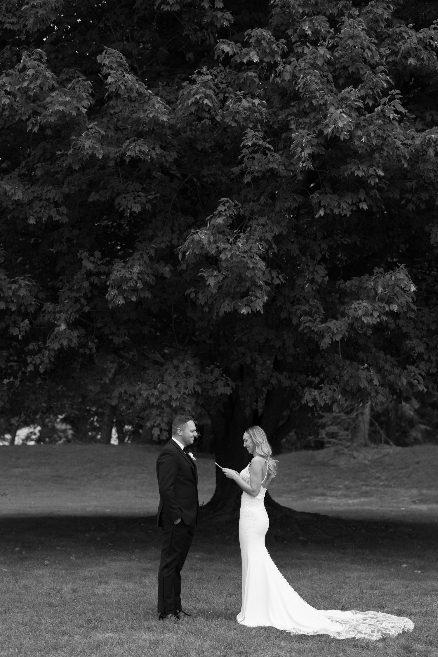 Bride reads vows to groom in black and white portrait beneath a large tree.
