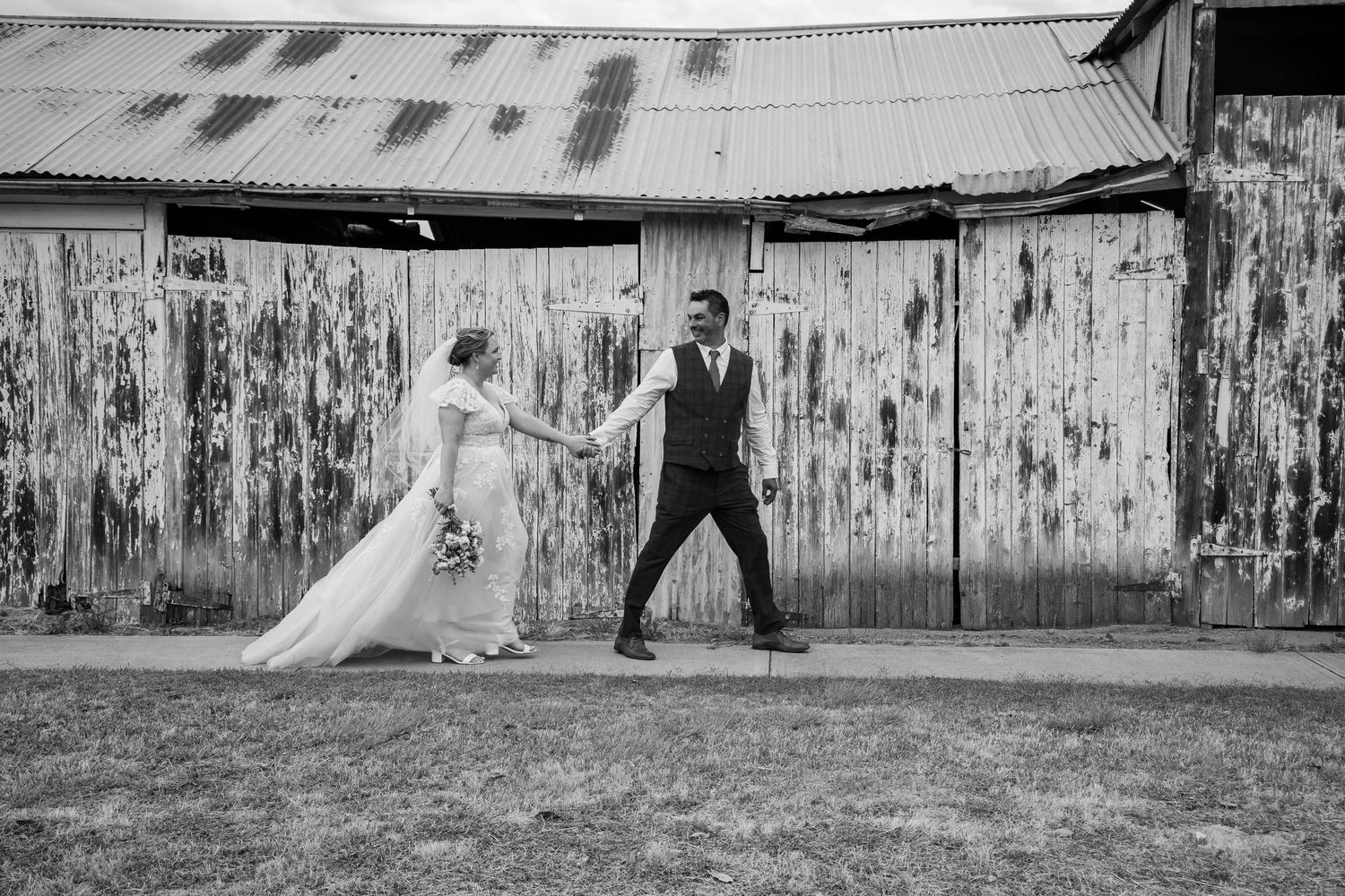 Black and white photo of a bride and groom walking along a sandy riverbank surrounded by eucalyptus trees.