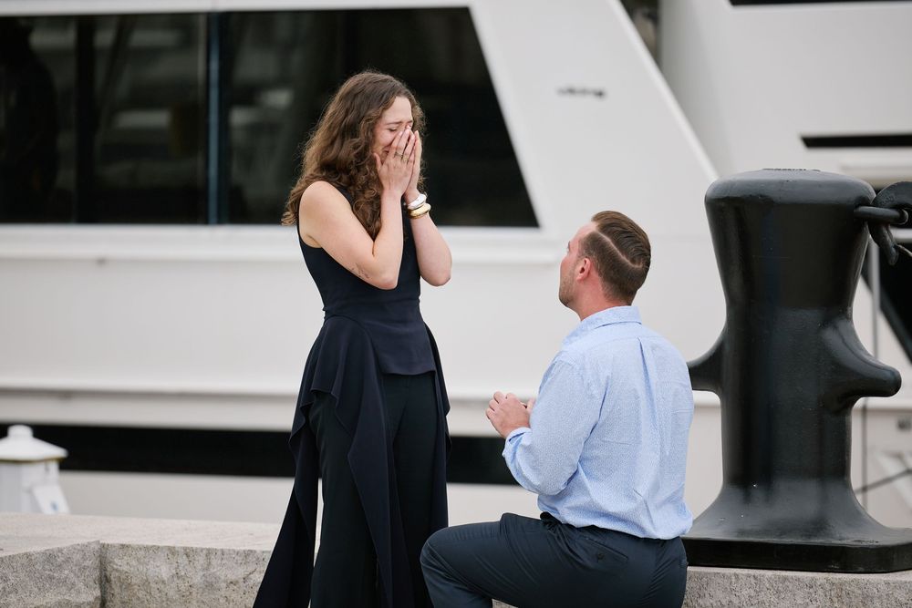 Man proposes on one knee to surprised woman covering her mouth near a marina with a yacht in the background.