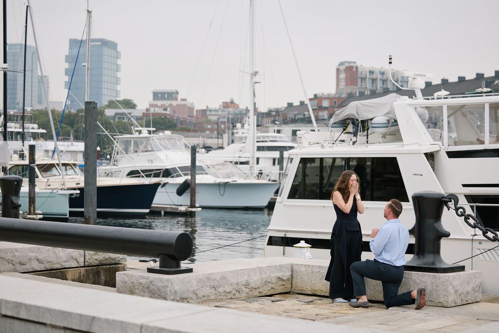 Man proposes to woman on a marina dock with yachts and Boston skyline in the background.