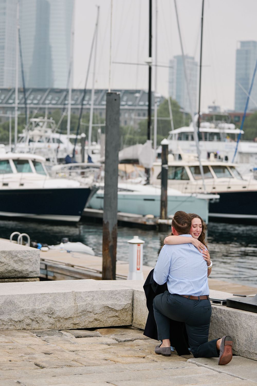 Man proposes to woman at a marina waterfront, with sailboats and city skyline in the background.
