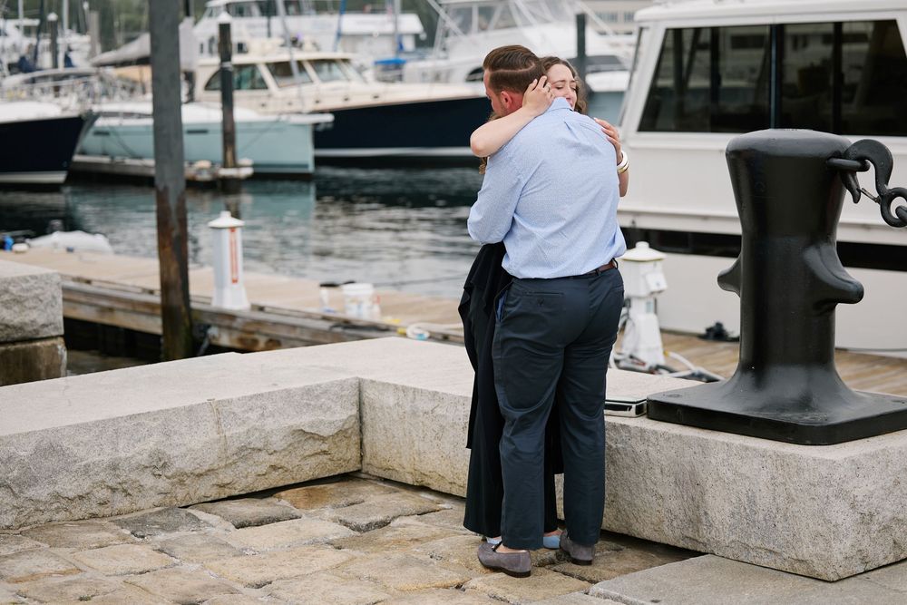 Couple embracing on a marina dock with boats and water in the background on a cloudy day.