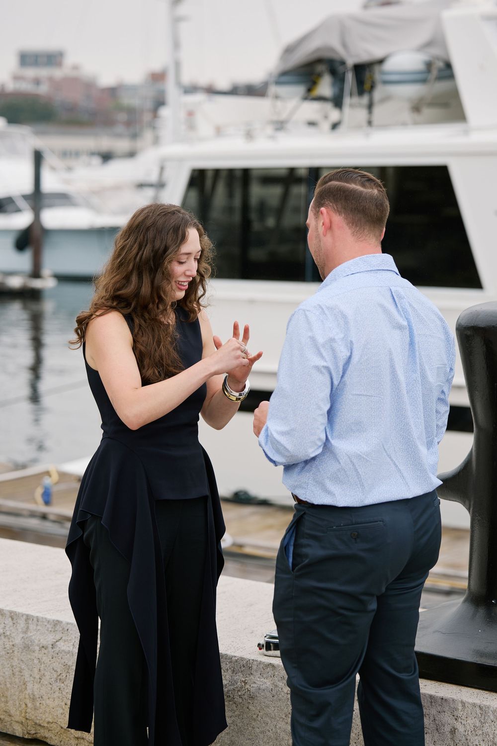 Man placing engagement ring on woman's finger at a marina with yachts in the background.