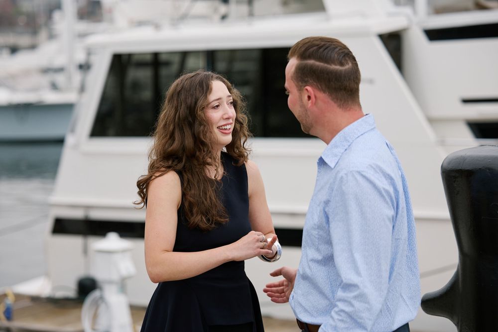 A smiling couple in elegant attire chat on a marina dock with luxury yachts in the background.