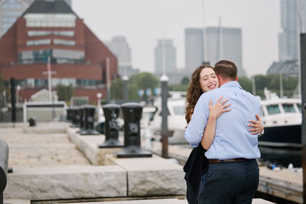 Couple embracing and smiling at a waterfront harbor with city skyline and boats in the background.